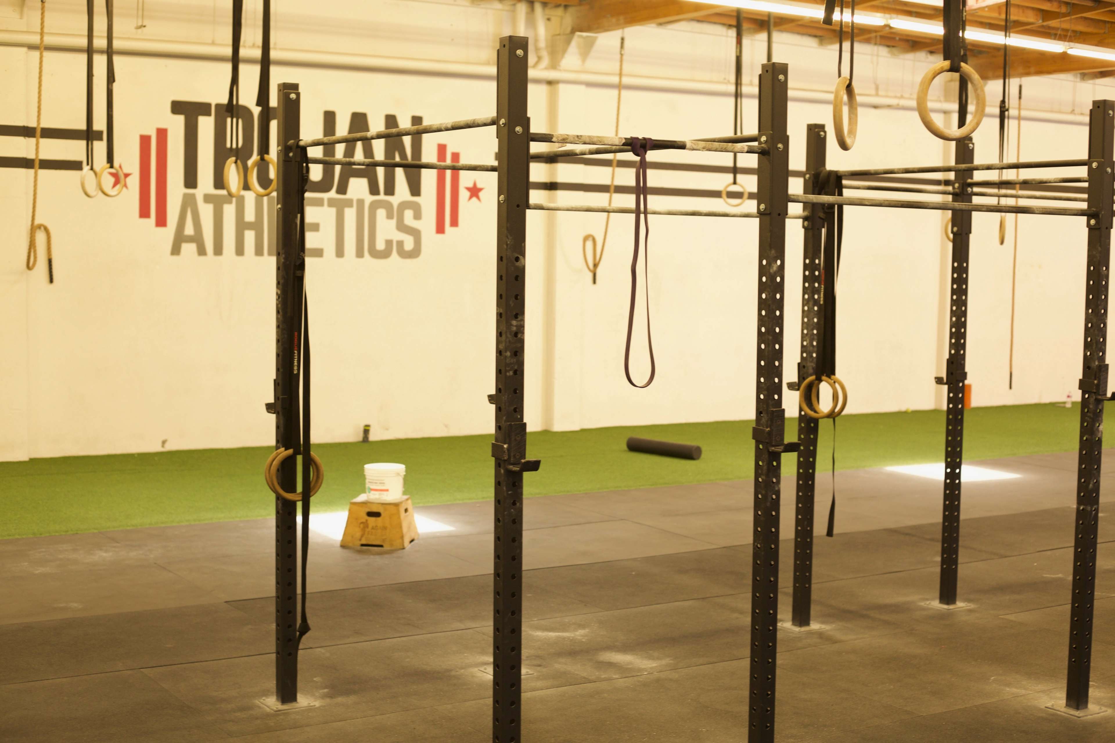 The image shows a gym interior with black workout equipment, including pull-up bars and rings, against a backdrop with the text "Trojan Athletics" on the wall.