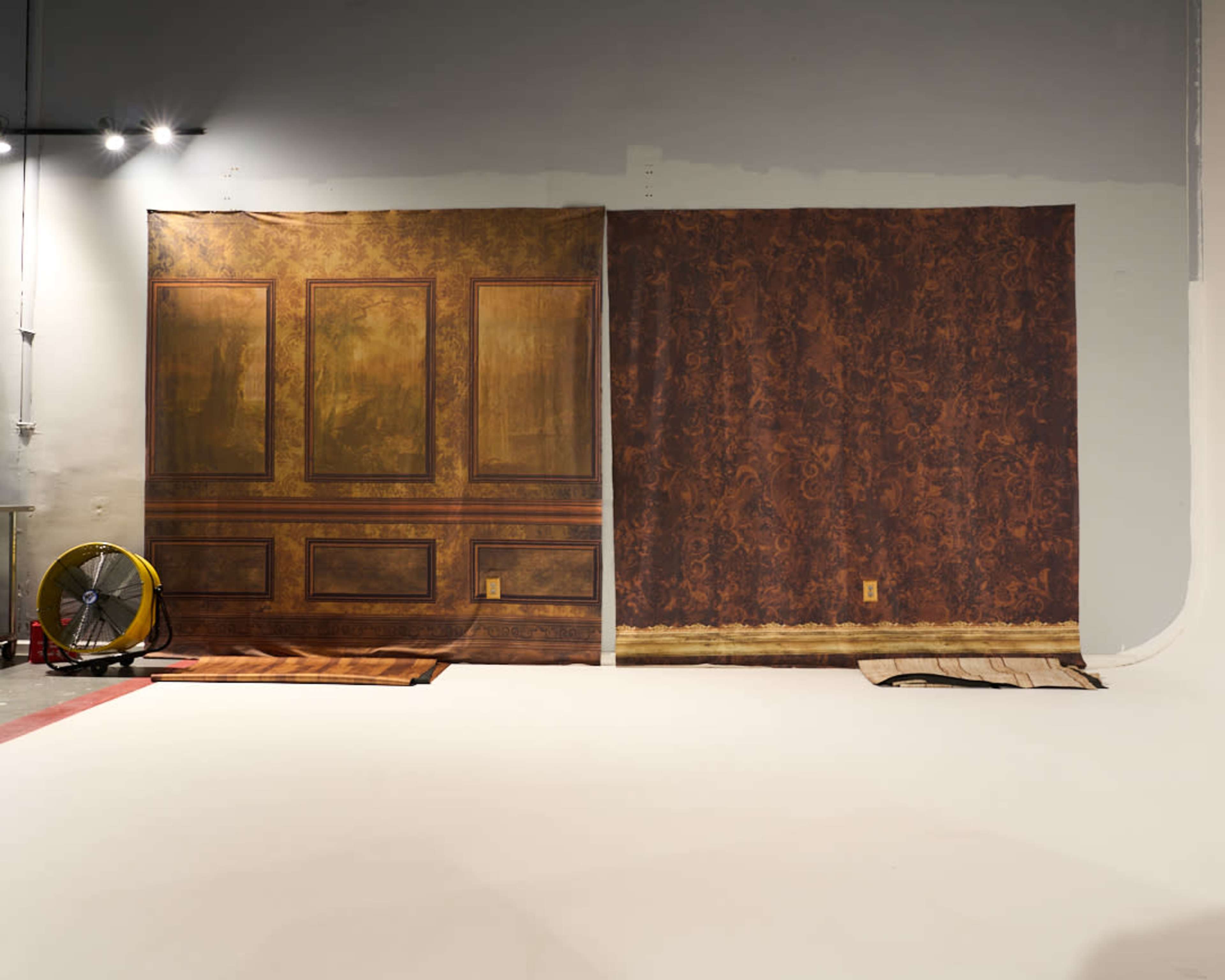 Two large backdrops against a gray wall, one resembling an ornate wooden paneling and the other depicting a textured brown wall, with a fan and some materials on the floor nearby.