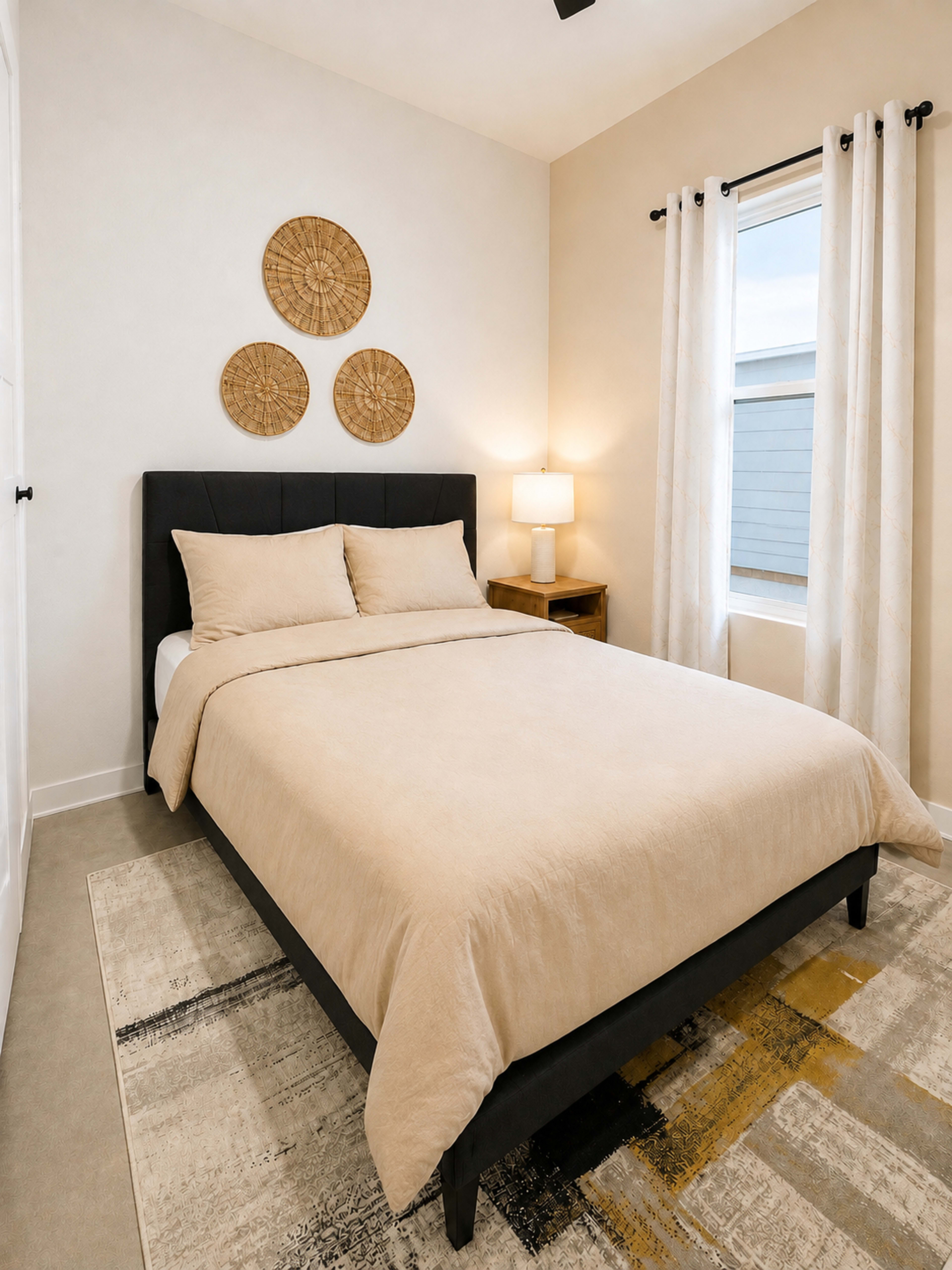 A neatly arranged bedroom features a bed with a beige duvet, decorative wall hangings, a bedside table with a lamp, and a large window partly covered by curtains.