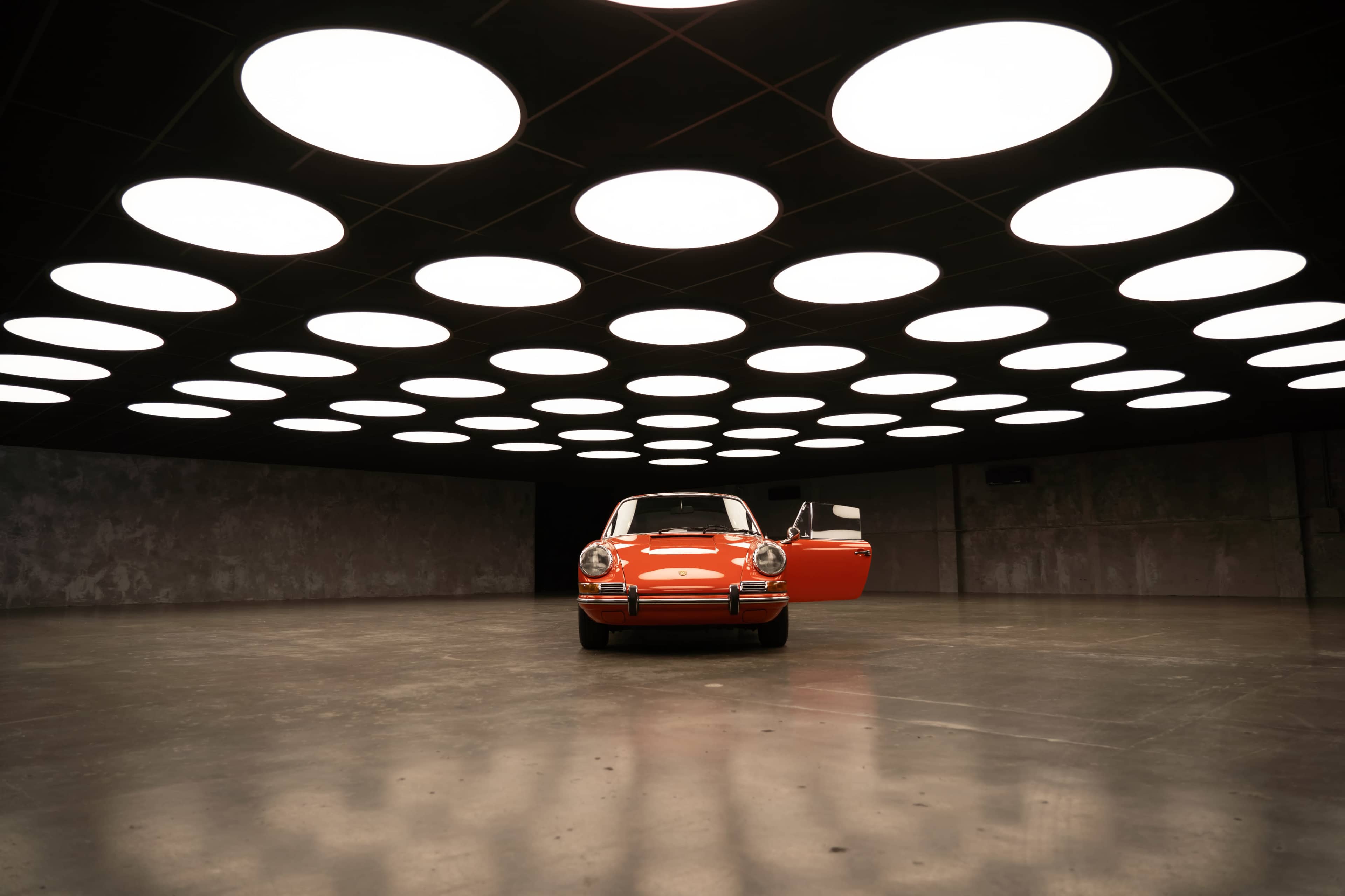 A vintage orange car is positioned in a spacious, dimly lit room with circular overhead lights.