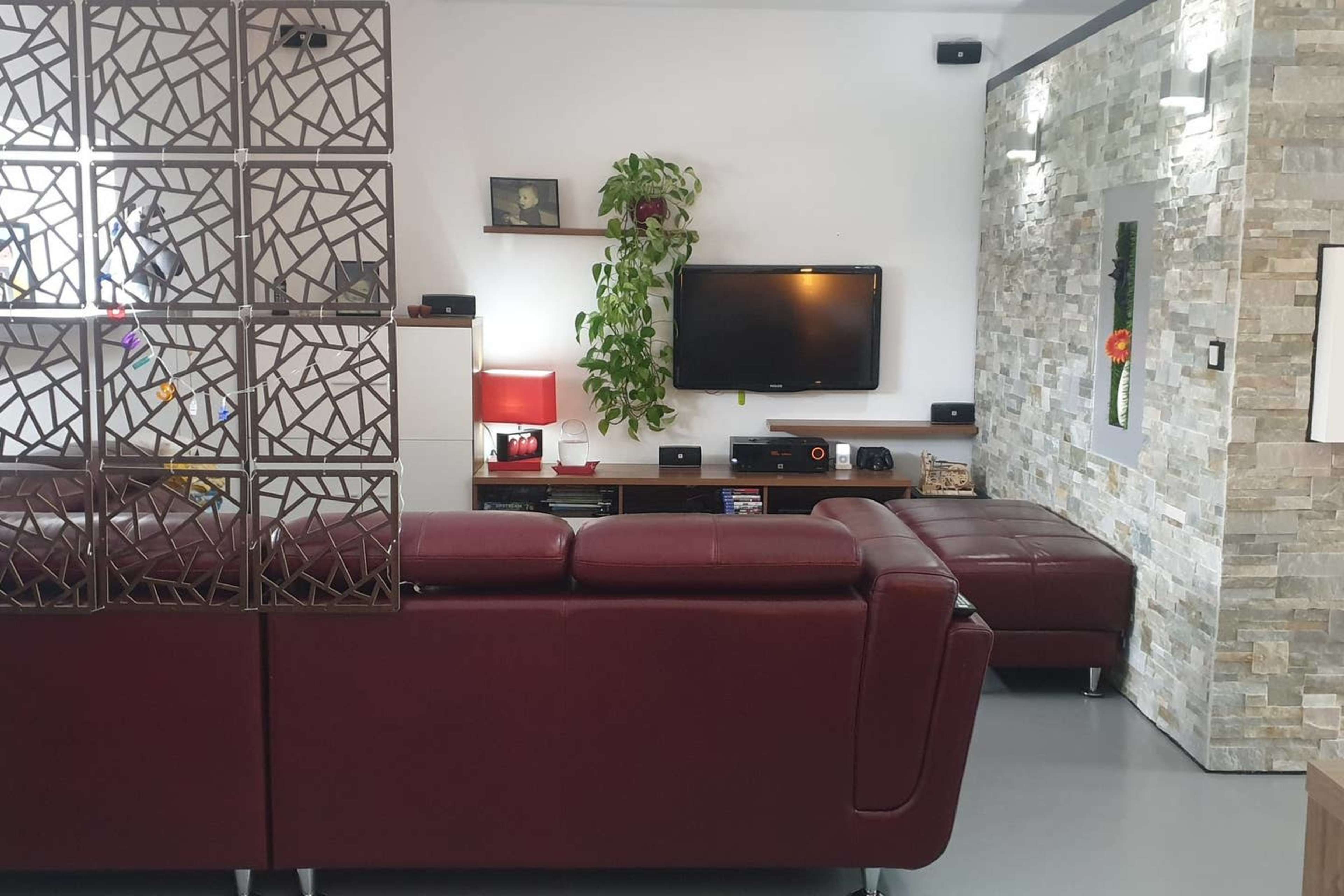 The image shows a modern living room featuring a maroon couch, a decorative partition, a wall-mounted television, and stone accent walls.