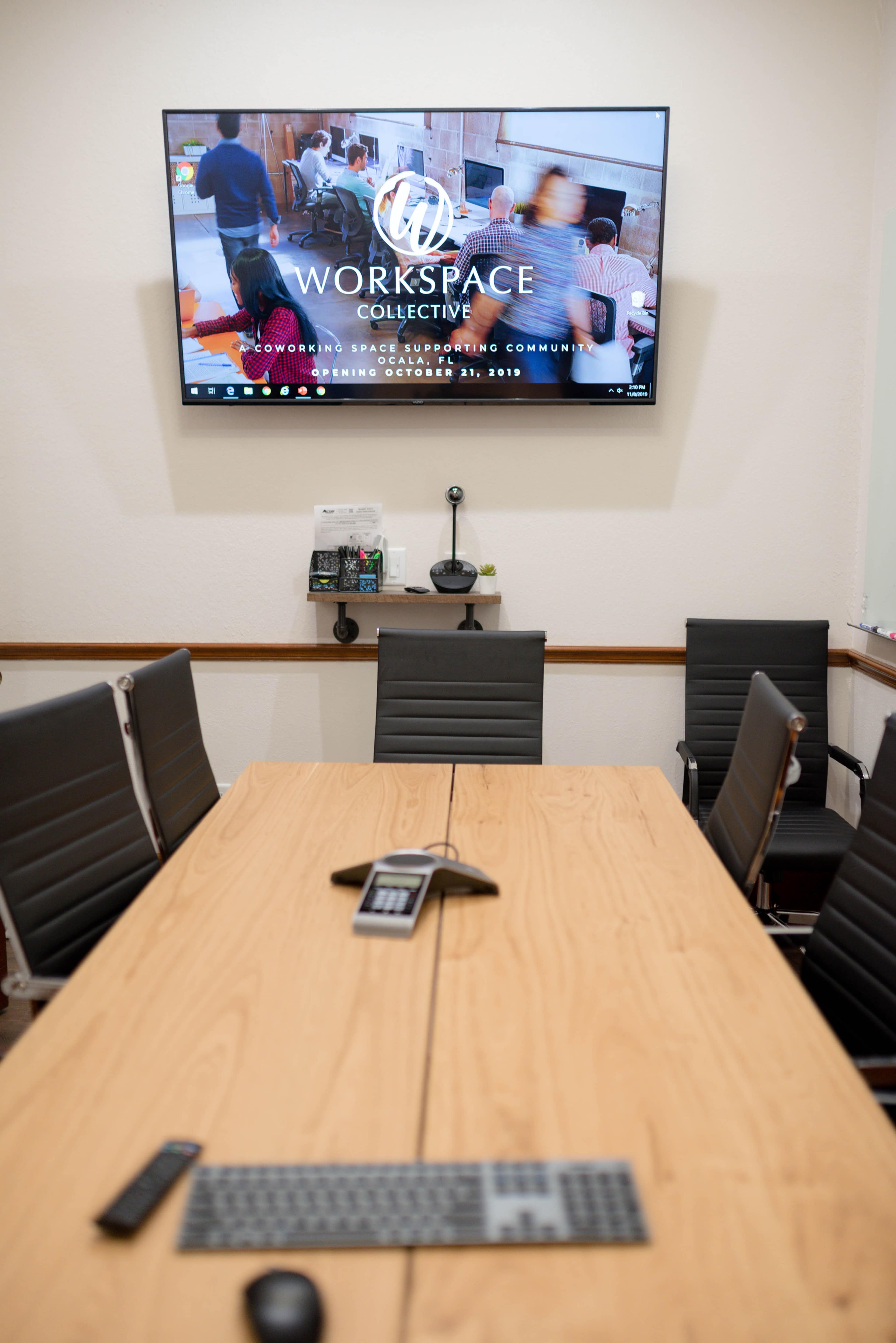A conference room features a wooden table surrounded by black chairs, with a large screen displaying the logo and information for "Workspace Collective" on the wall.