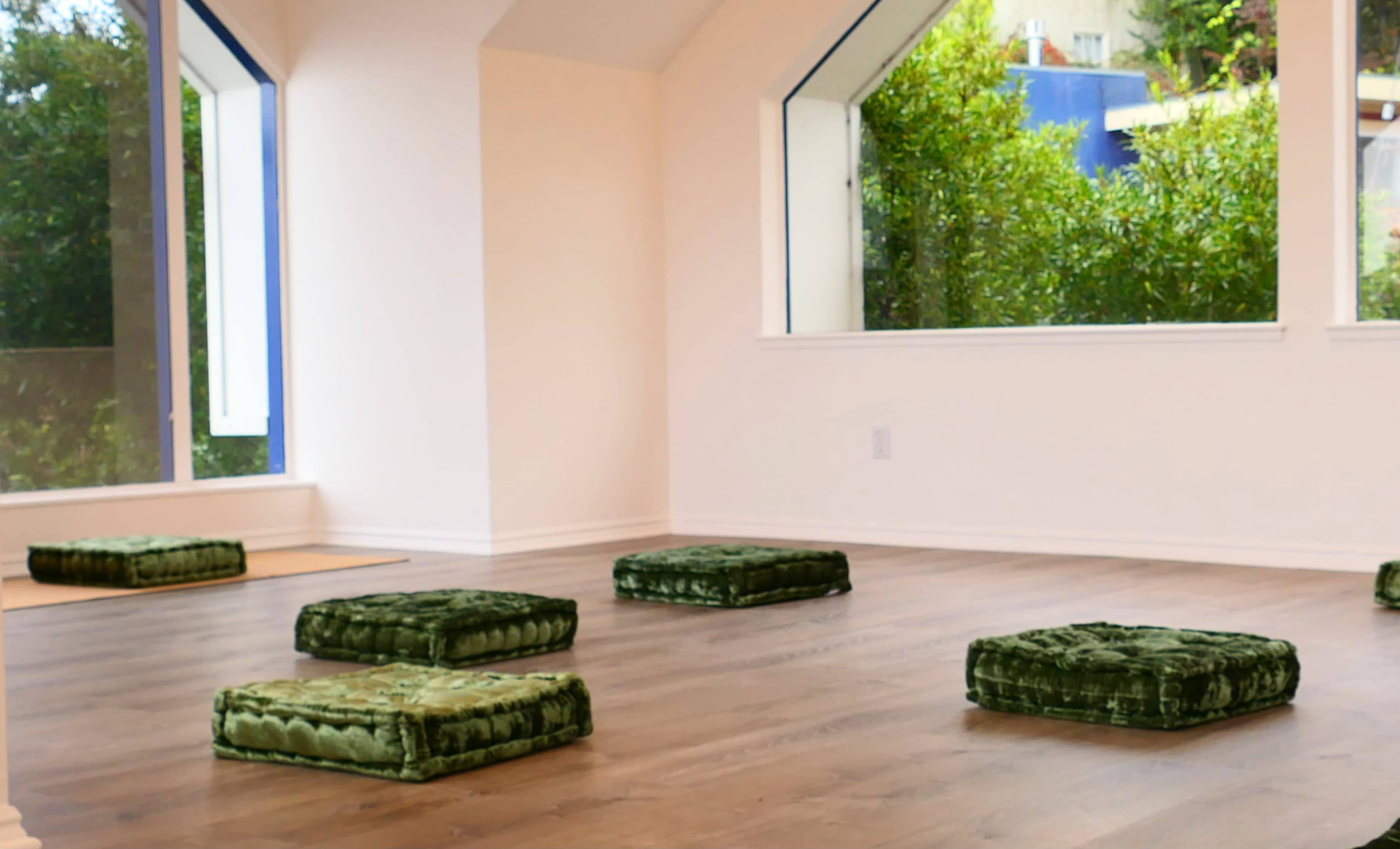 A room with a wooden floor and several green meditation cushions arranged on it, surrounded by large windows that provide natural light and a view of greenery outside.