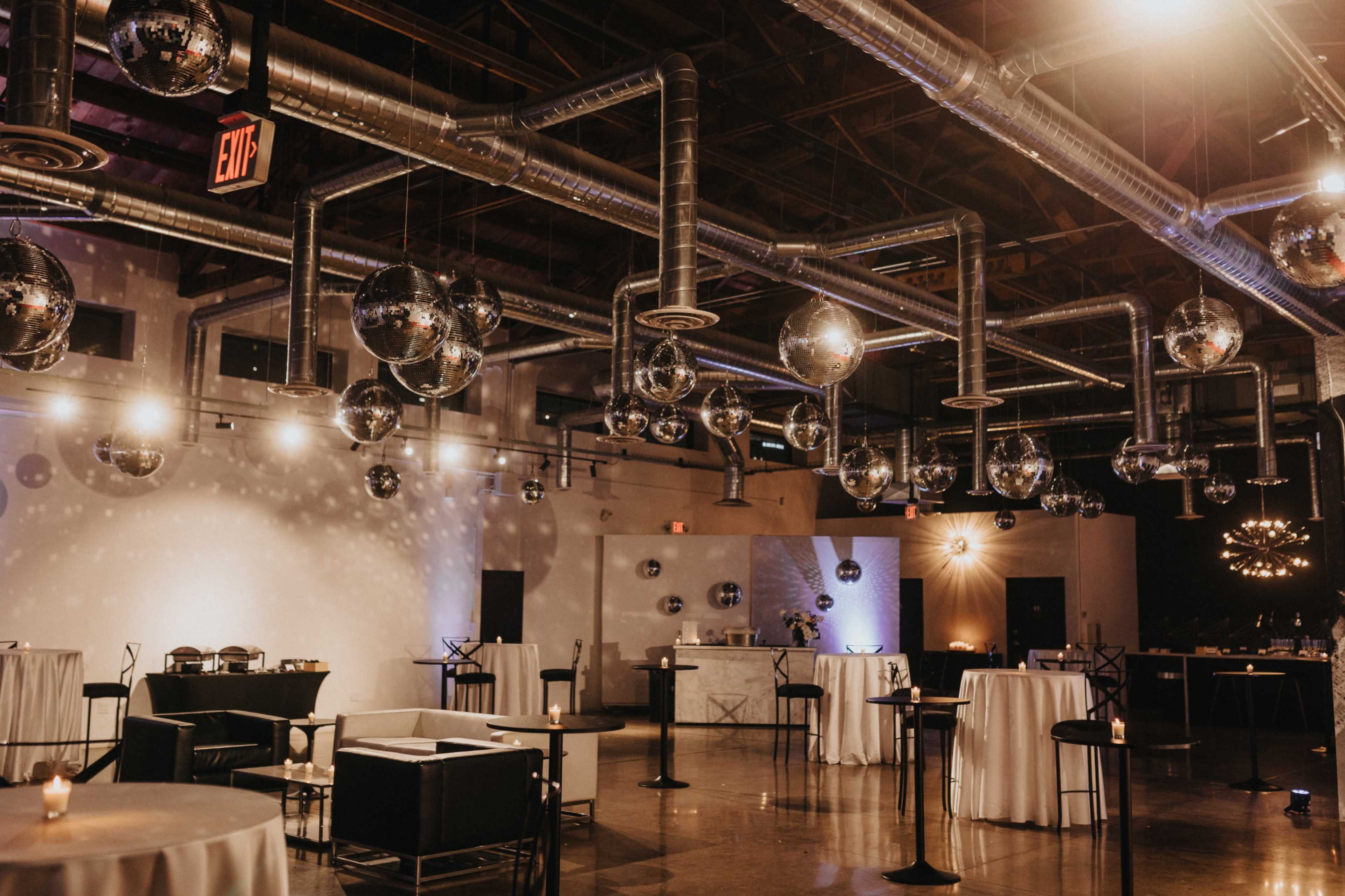 A spacious event venue features circular tables, black seating, and hanging disco balls reflecting light from overhead fixtures.