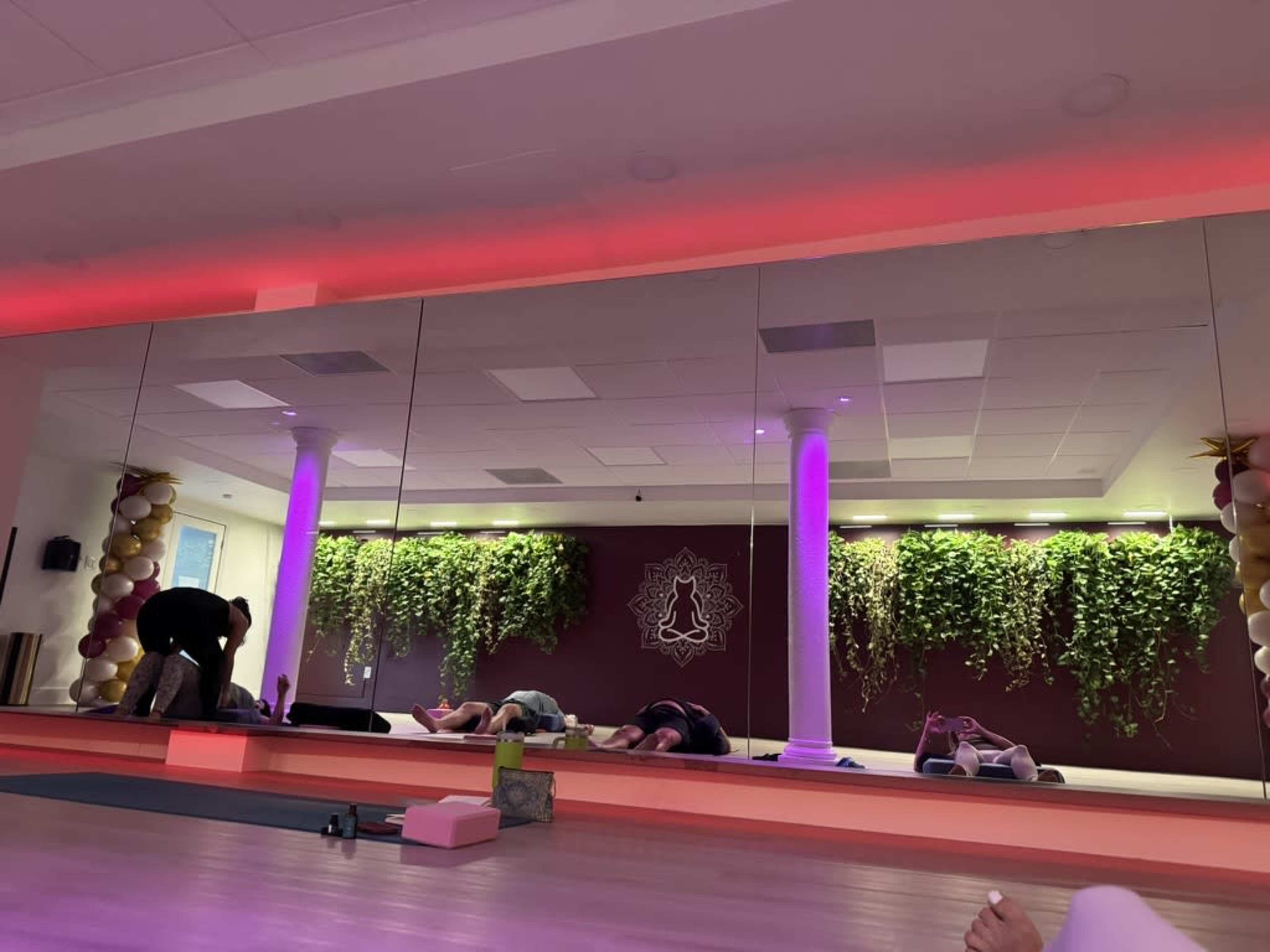 The image shows a yoga studio with a mirrored wall, featuring plants and colored lighting, where individuals are practicing yoga on mats.