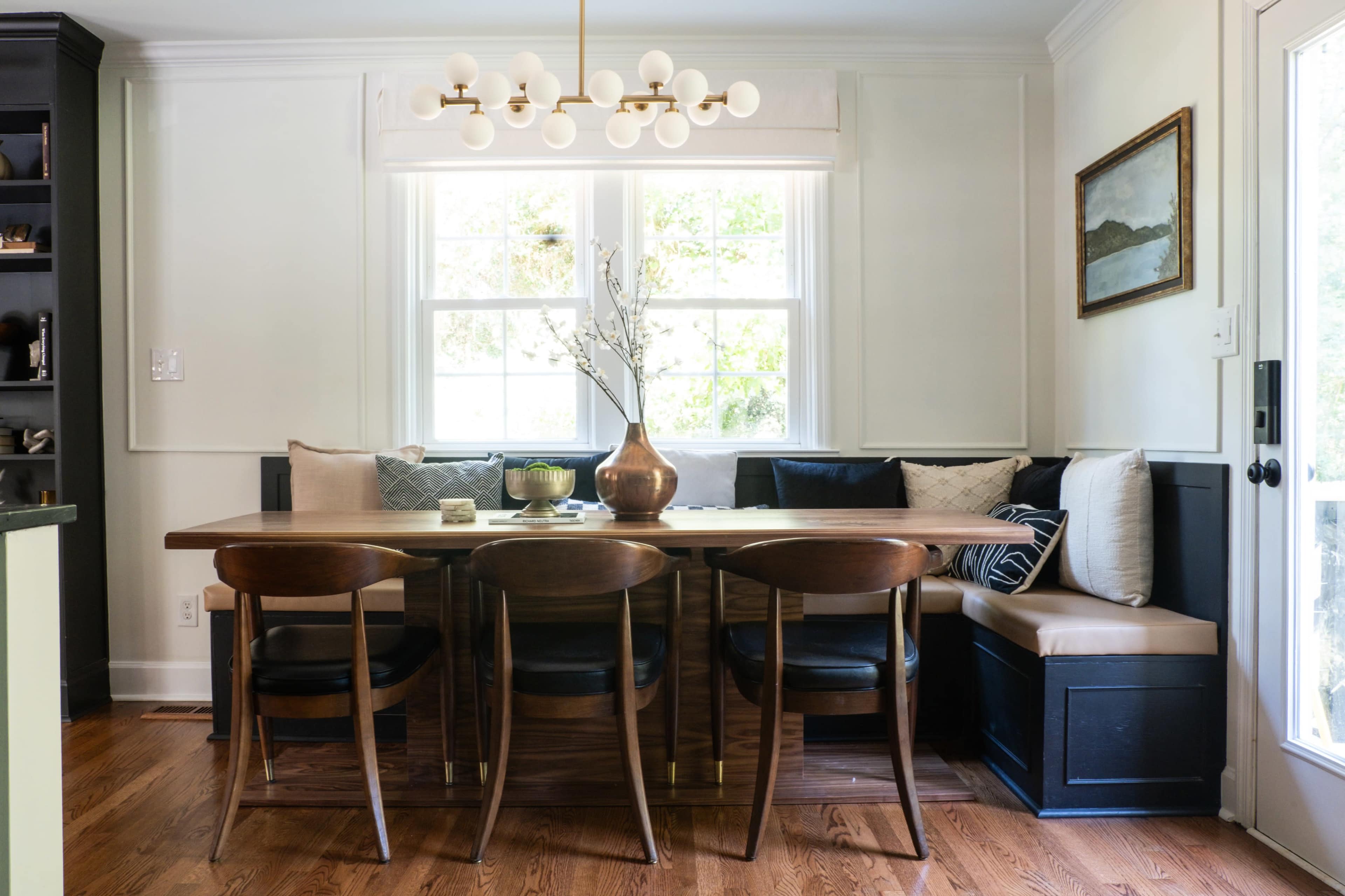A wooden dining table surrounded by chairs is positioned near a window with a built-in bench seating area adorned with cushions.