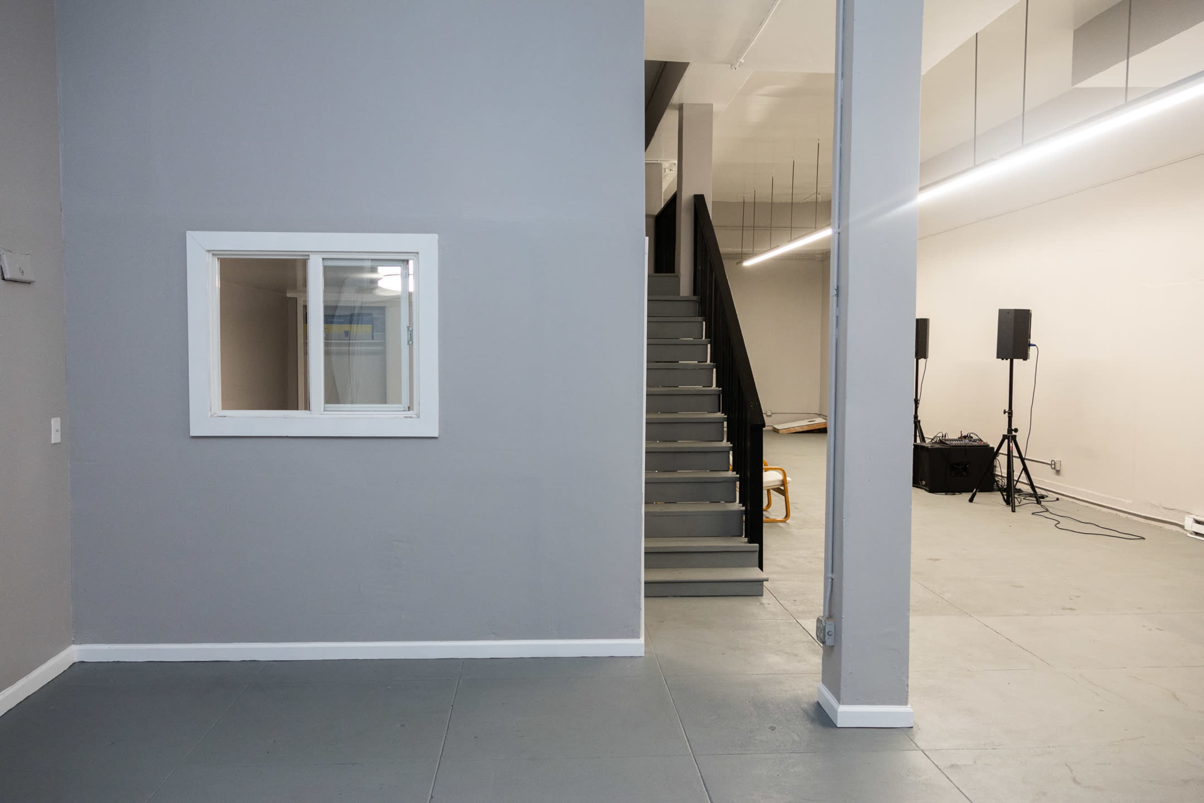 The image shows a modern interior space featuring a gray wall with a window, a staircase leading to an upper level, and an empty area with sound equipment on the floor.