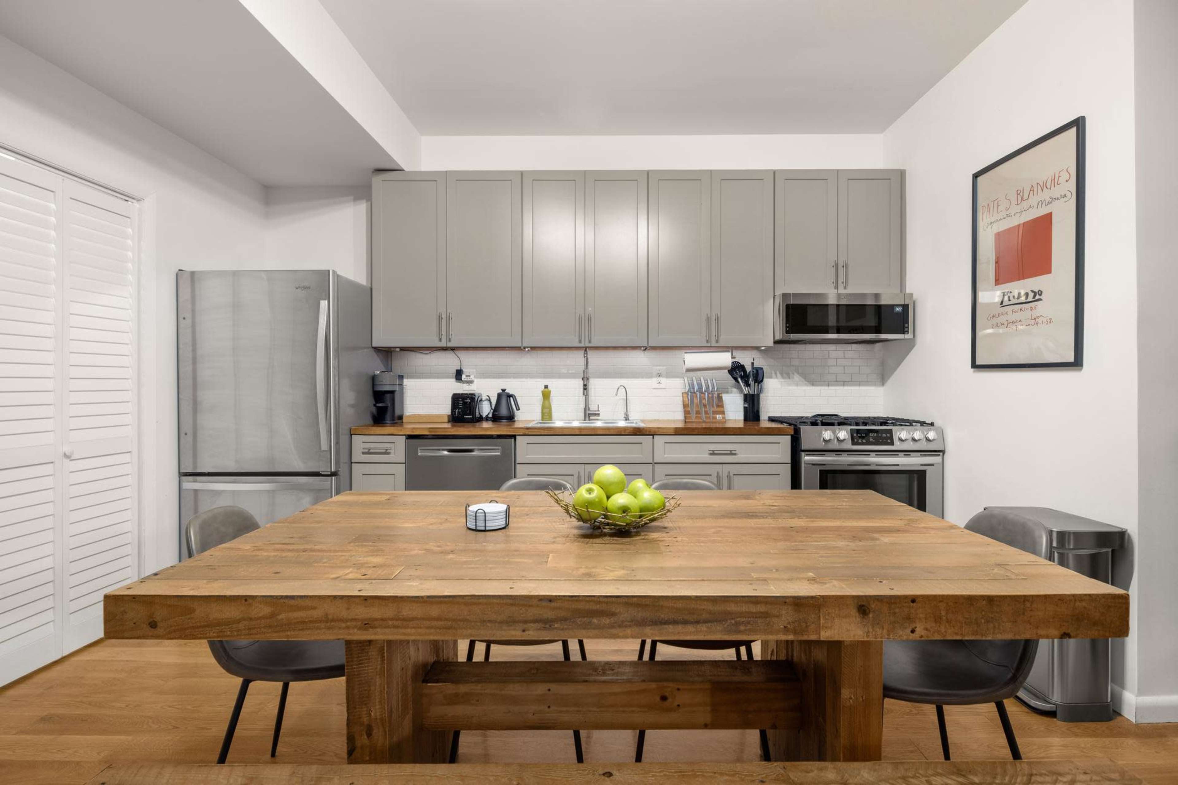 A modern kitchen with gray cabinets, stainless steel appliances, and a large wooden dining table set with a bowl of green apples.