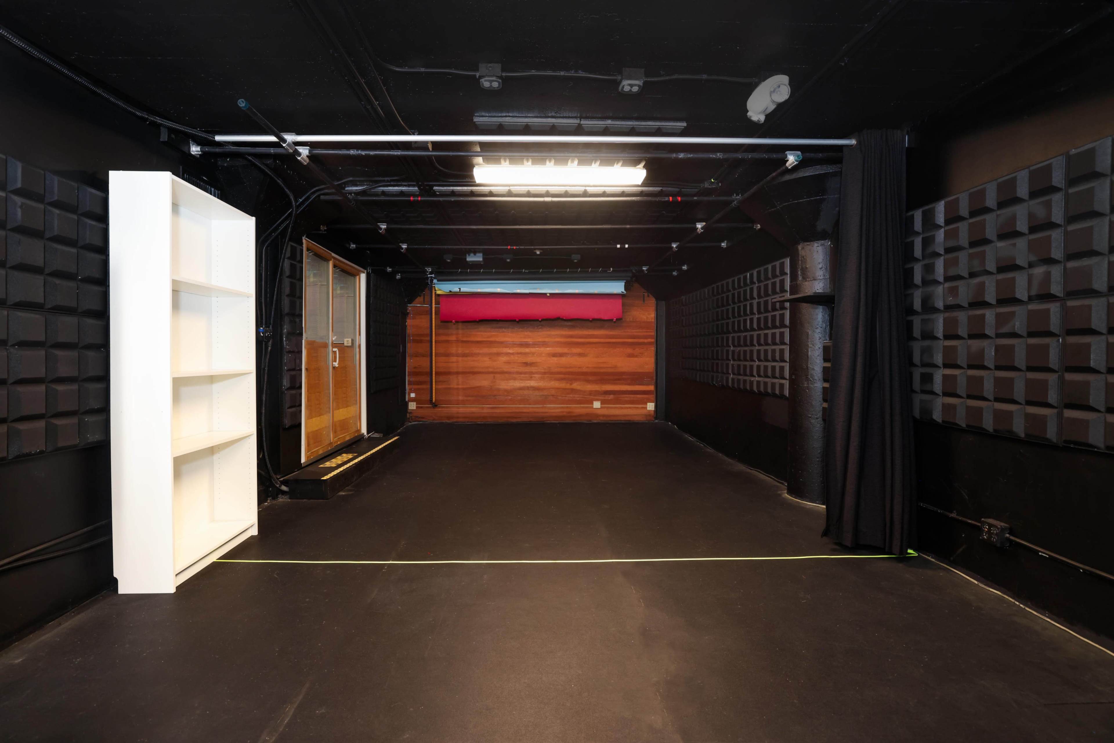 The image shows a large, dark studio space with black walls, a wooden panel at the back, and a white shelving unit on one side.