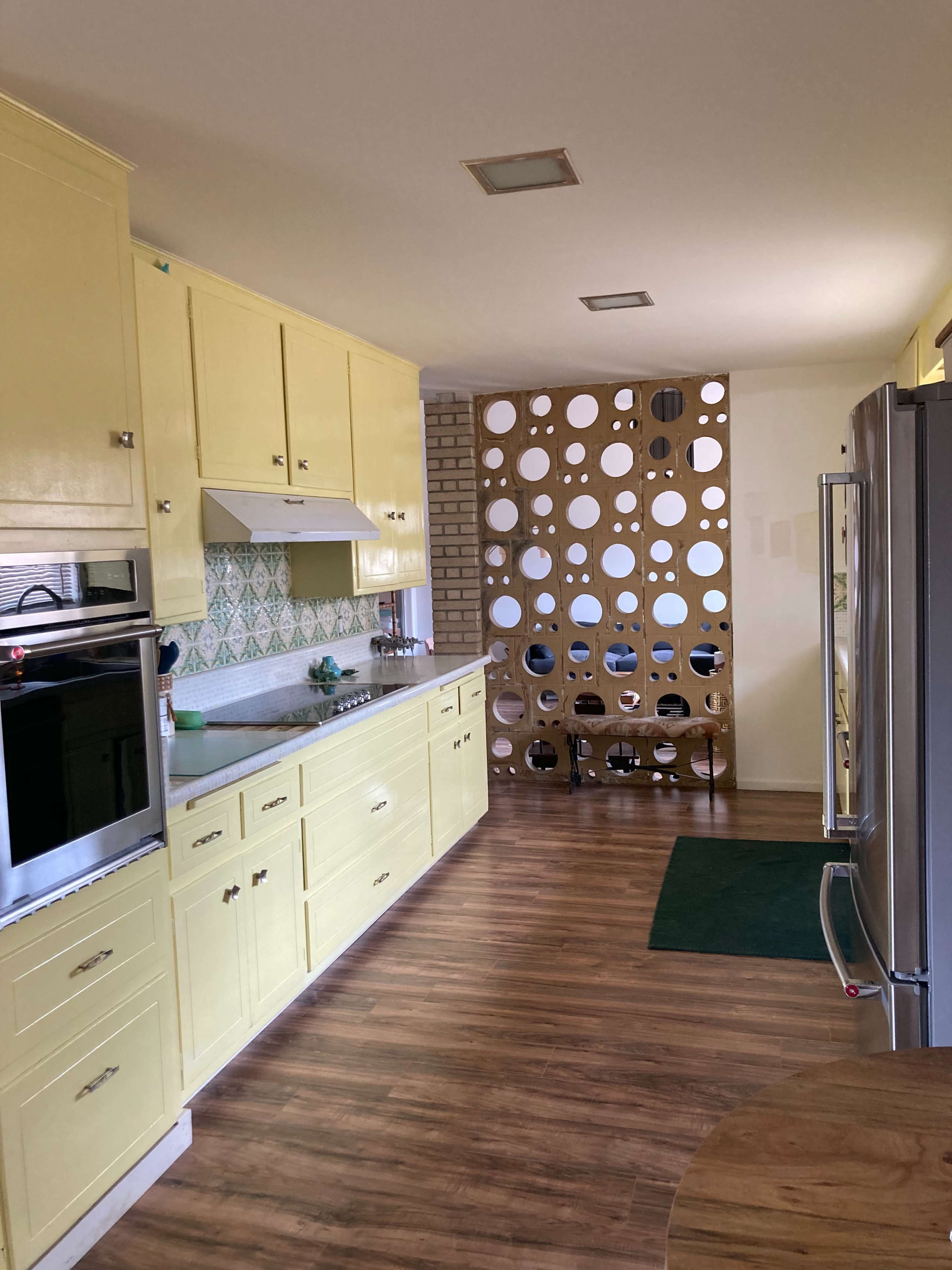 A kitchen features yellow cabinets, a tiled backsplash, and a wooden divider with circular cutouts separating it from a living area.