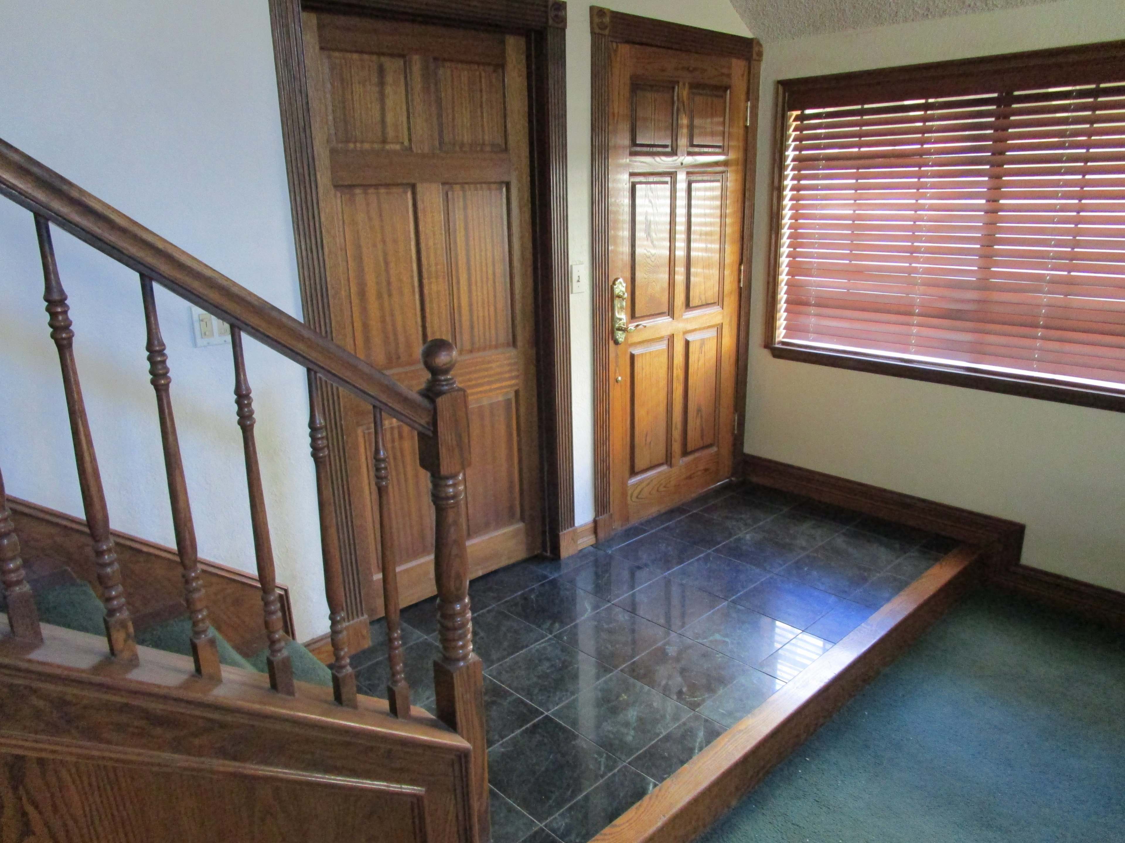 The image shows a hallway with wooden doors and a bench area, featuring a staircase, tile flooring, and a window with wooden blinds.