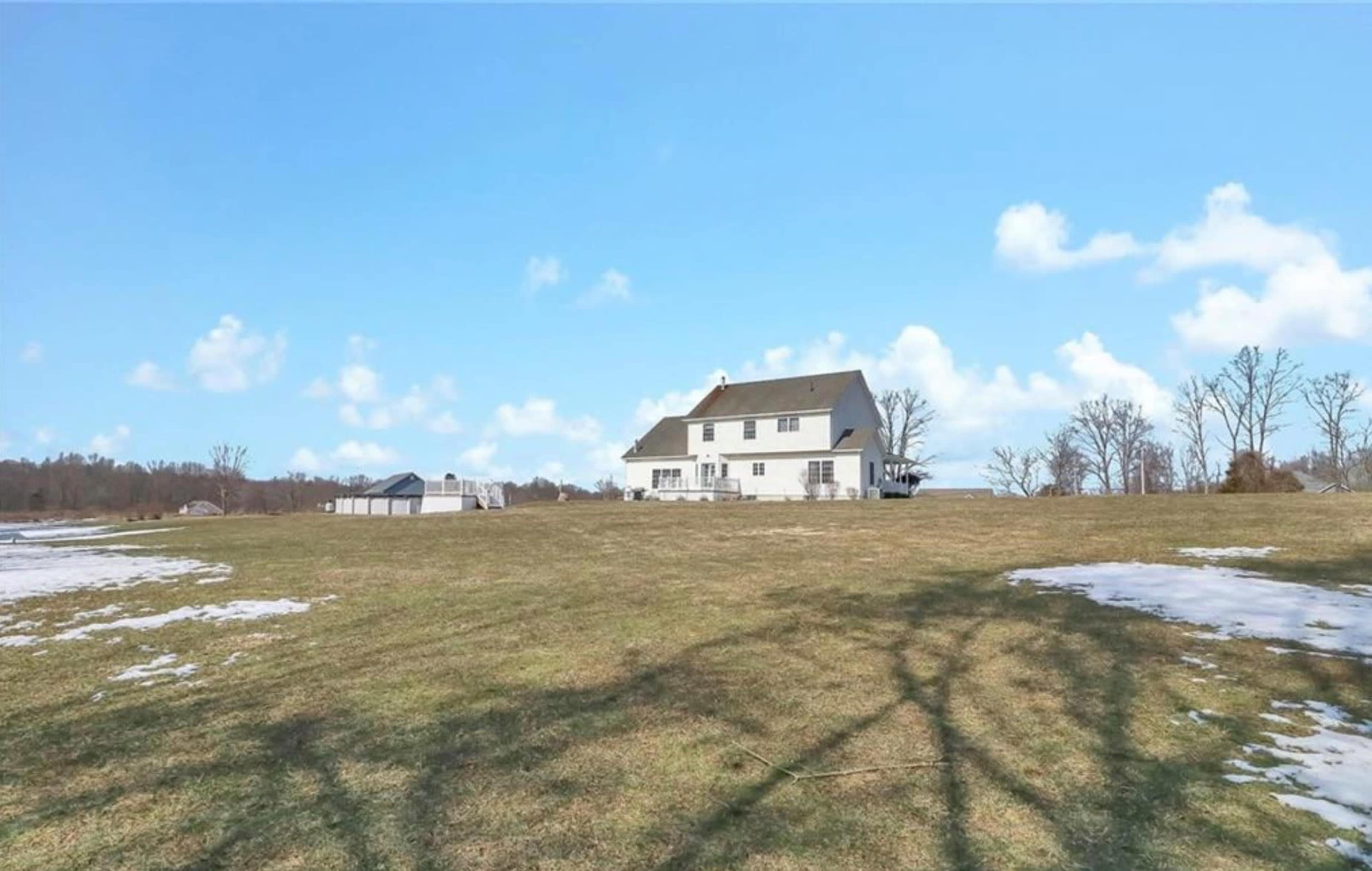 A white two-story house is set in a spacious grassy field with patches of snow and a clear blue sky.