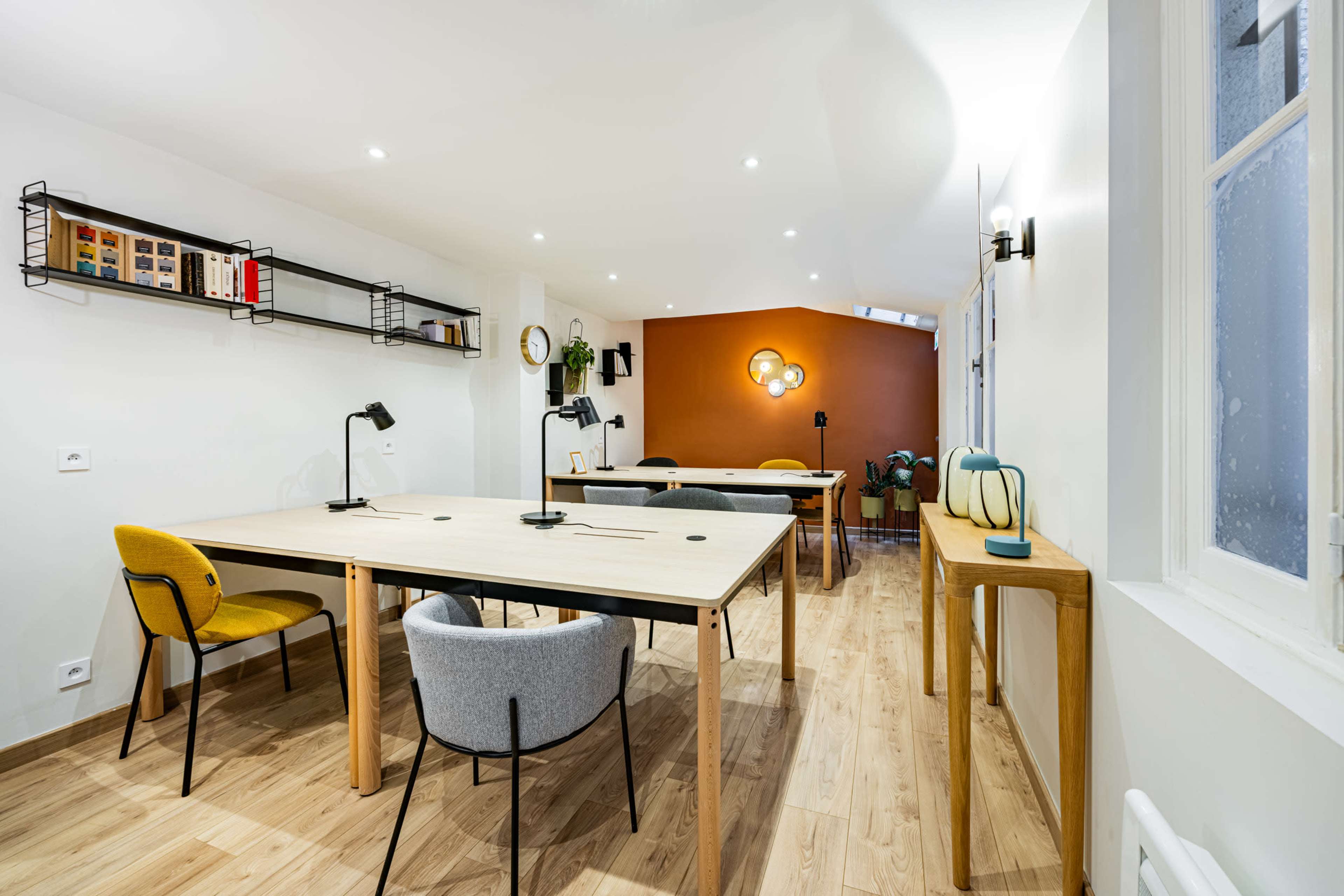 The image shows a modern office space with wooden floors, a large table in the center, and several task lights surrounding it, along with a feature wall painted in an orange hue.