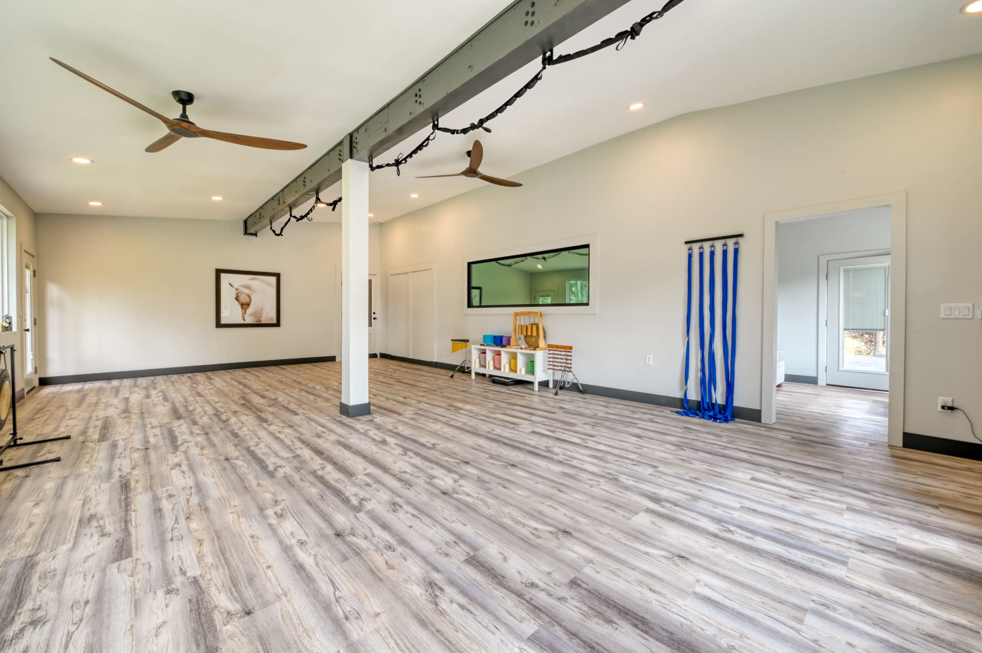 The image shows a spacious room with wooden flooring, large windows, two ceiling fans, and a divider that contains colorful drapes.
