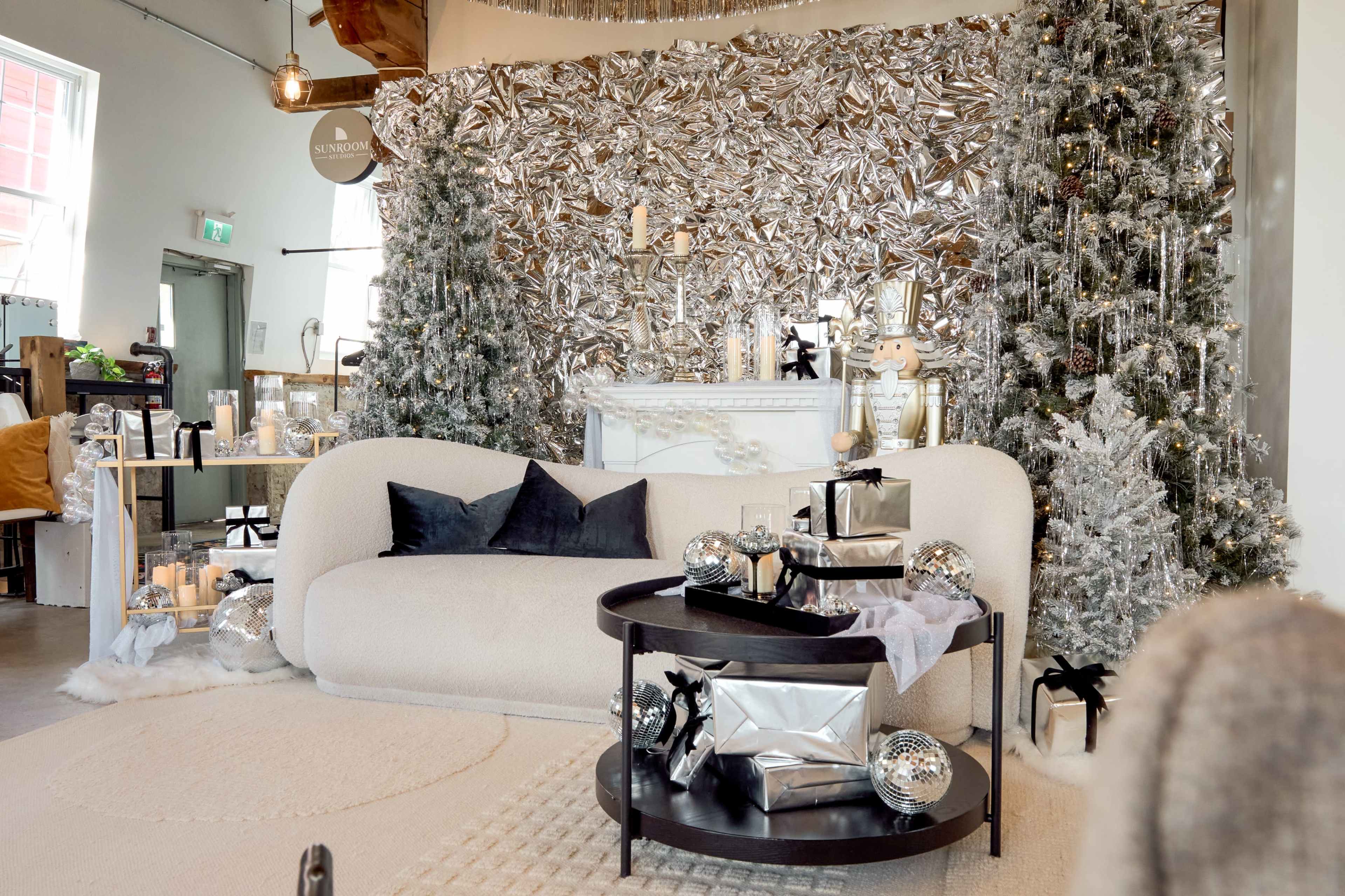 The image shows a modern living space decorated for the holidays, featuring a silver-themed backdrop, a plush sofa, and a coffee table adorned with wrapped gifts and decorative items.