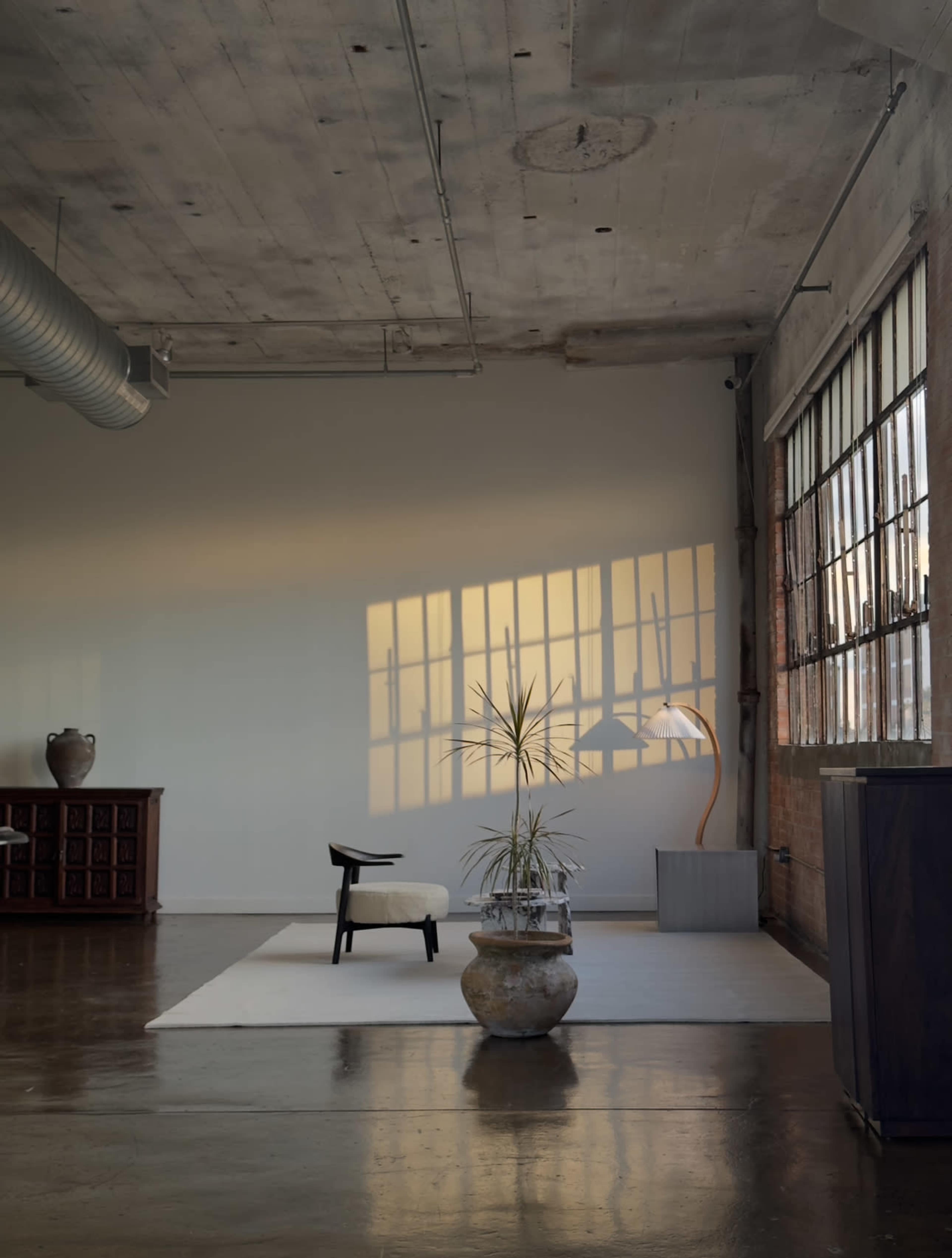 A minimalist interior features a chair, a potted plant, and a lamp against a backdrop of large windows with shadows on the wall.