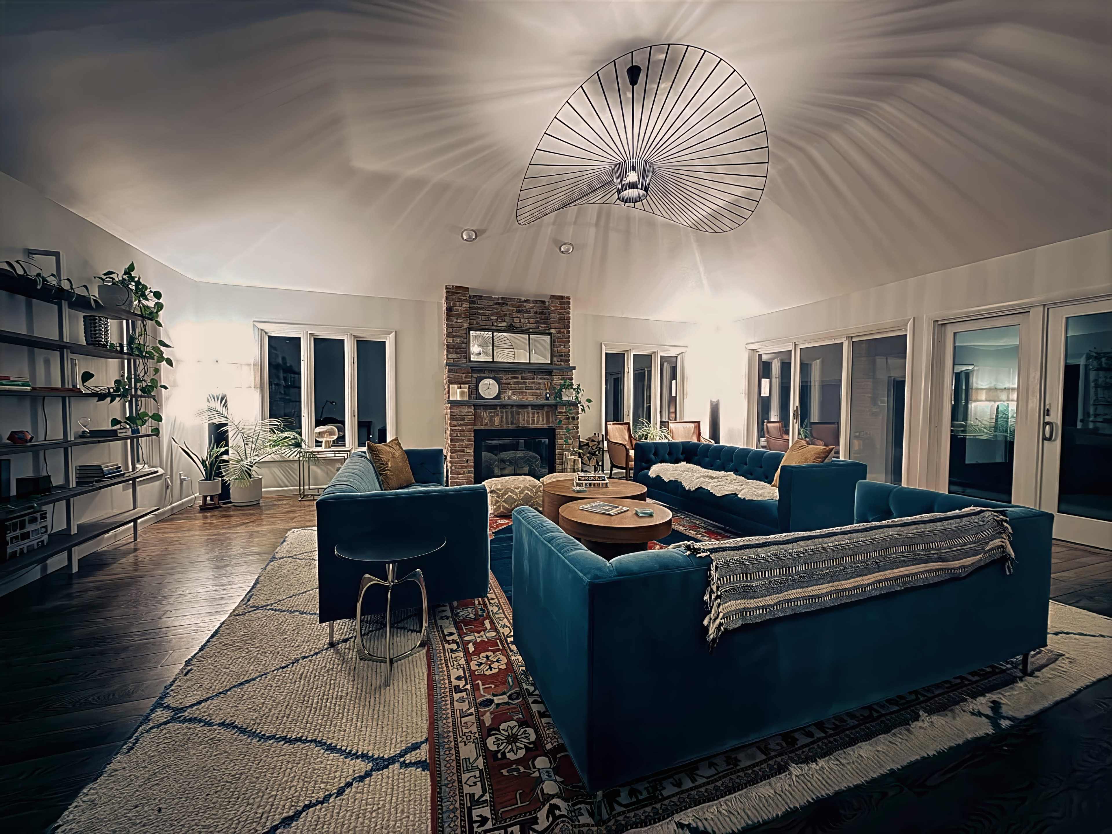 The image shows a spacious living room with a circular ceiling, featuring a large blue sectional sofa, a coffee table, and a brick fireplace surrounded by large windows.