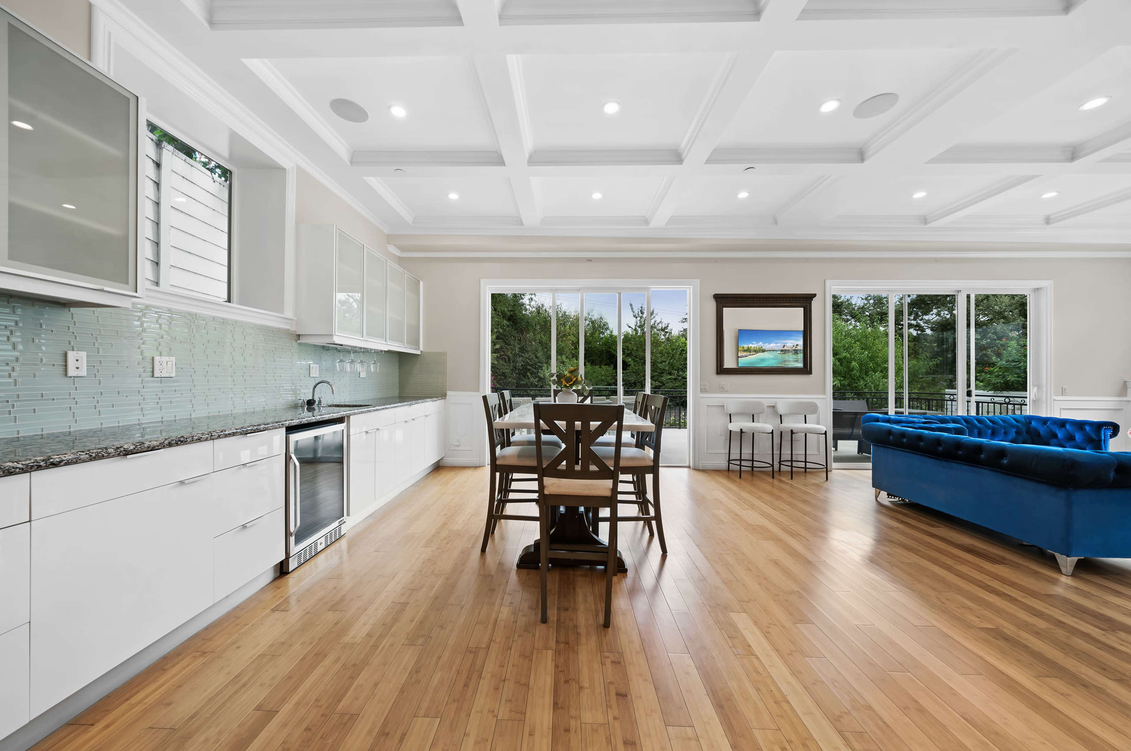 The image shows a modern, open-concept kitchen and dining area with a wooden dining table, a blue sofa, and large windows overlooking a green outdoor space.