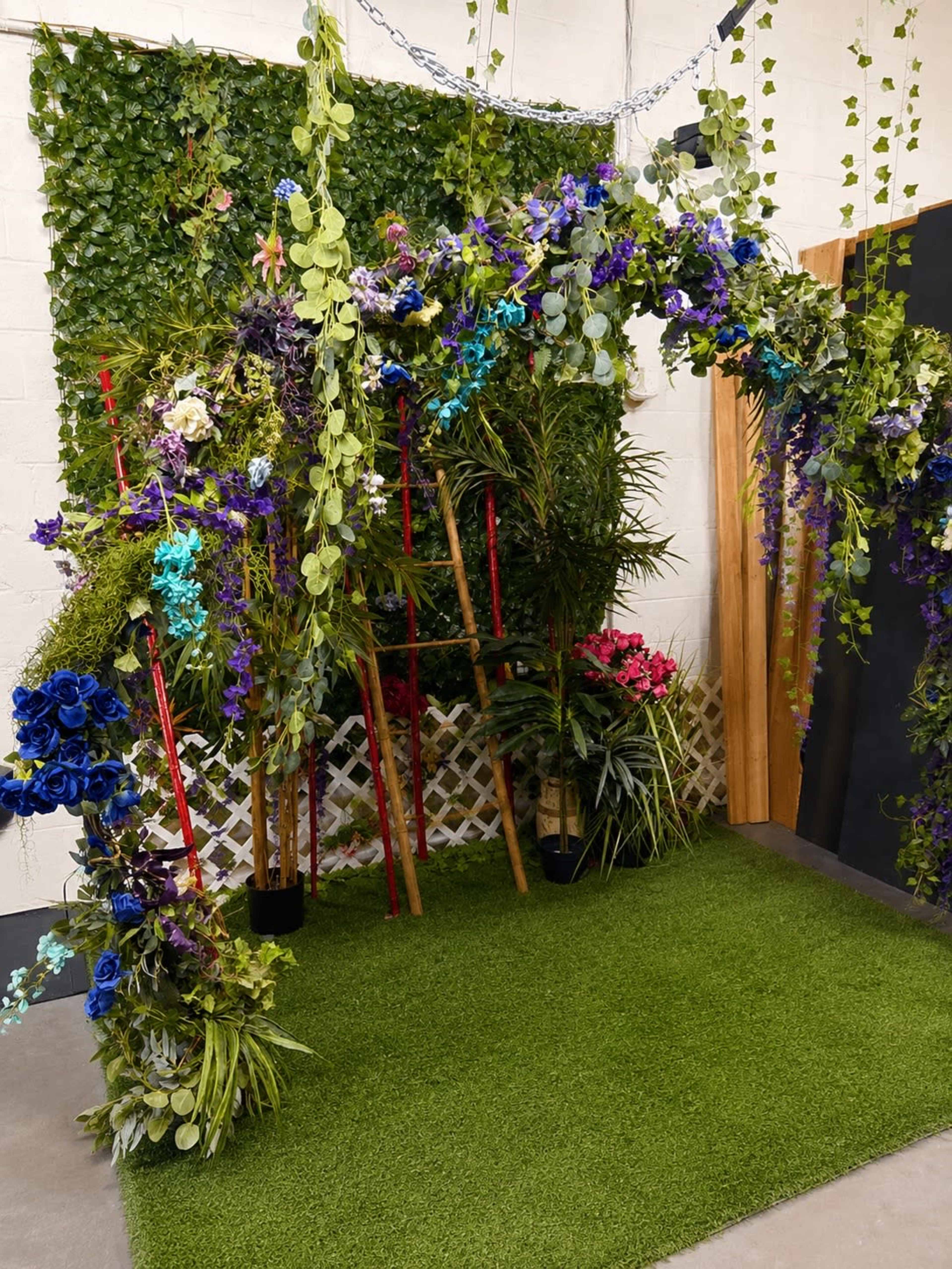 The image shows a floral arch decorated with various plants and flowers, set against a green backdrop in an indoor space.