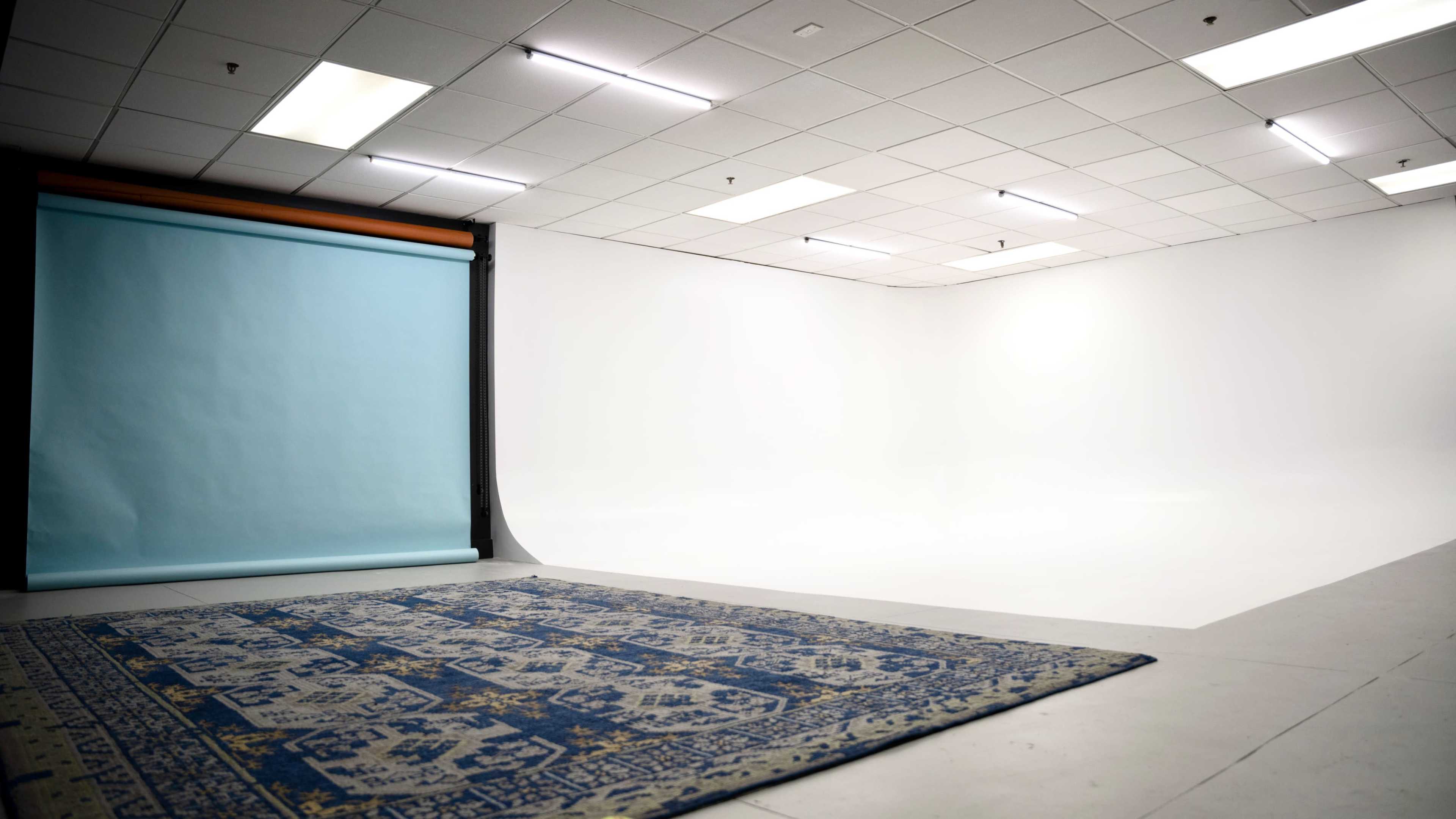 The image shows a photography studio featuring a blue backdrop, a large white seamless background, and a patterned rug on the floor.