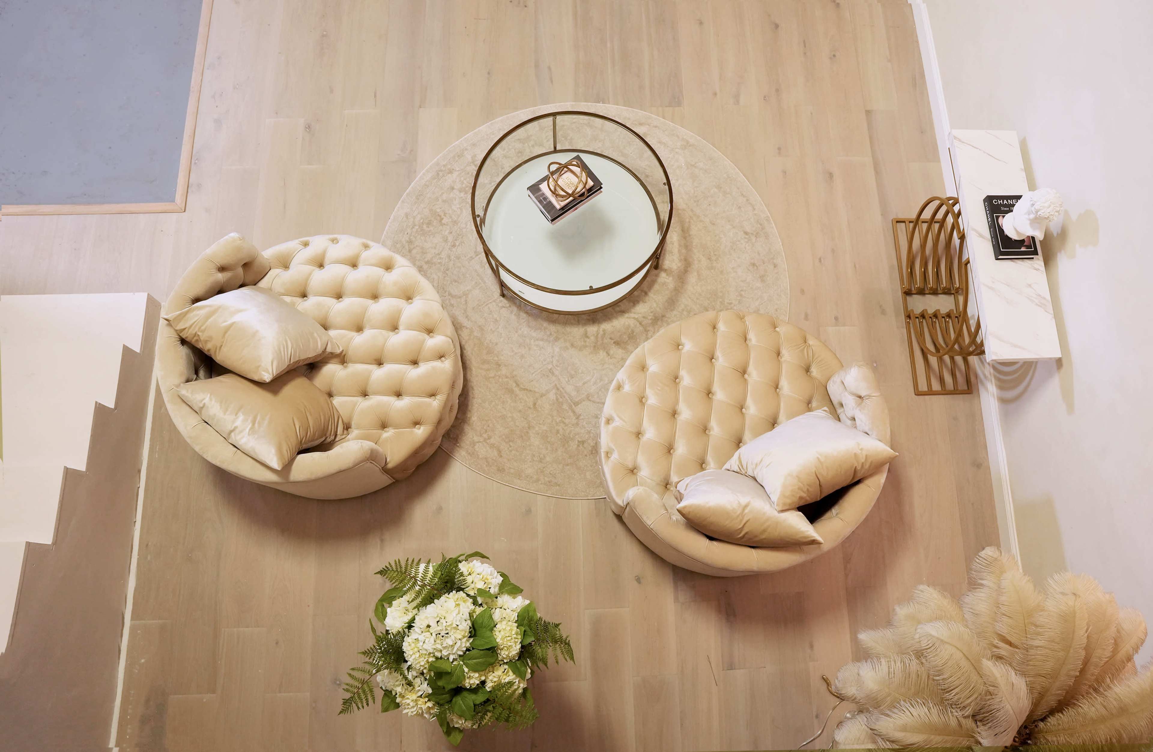 A modern living area with two tufted circular chairs facing a glass-top coffee table on a large round rug, accompanied by a potted plant and decorative items on a side table.