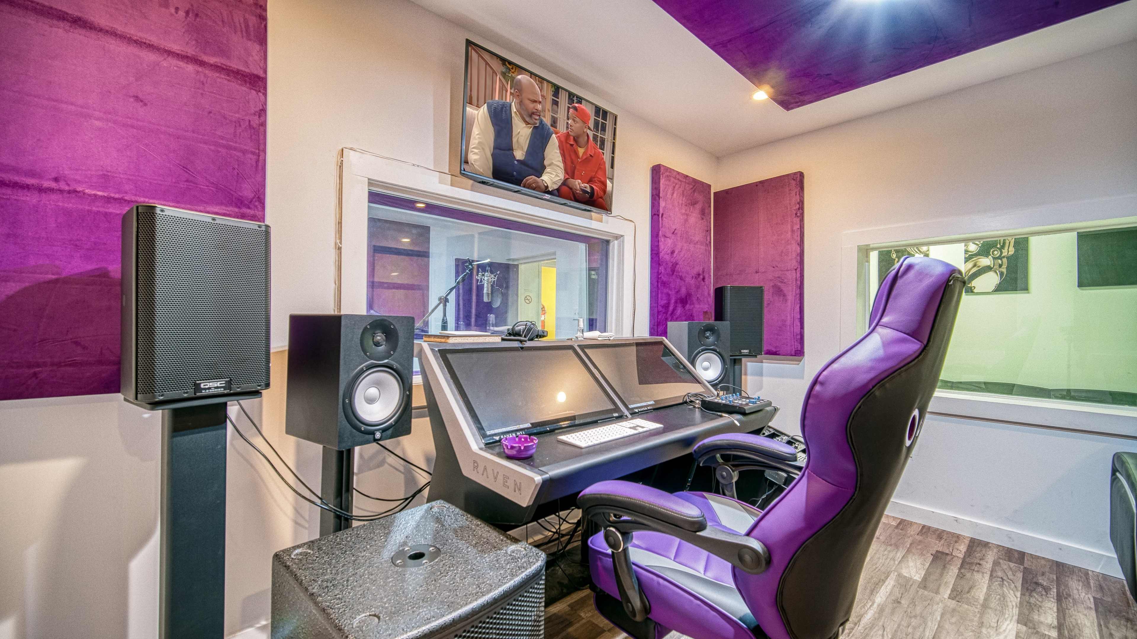 A recording studio with a control desk featuring speakers, a computer monitor displaying a TV show, and purple soundproofing panels on the walls.