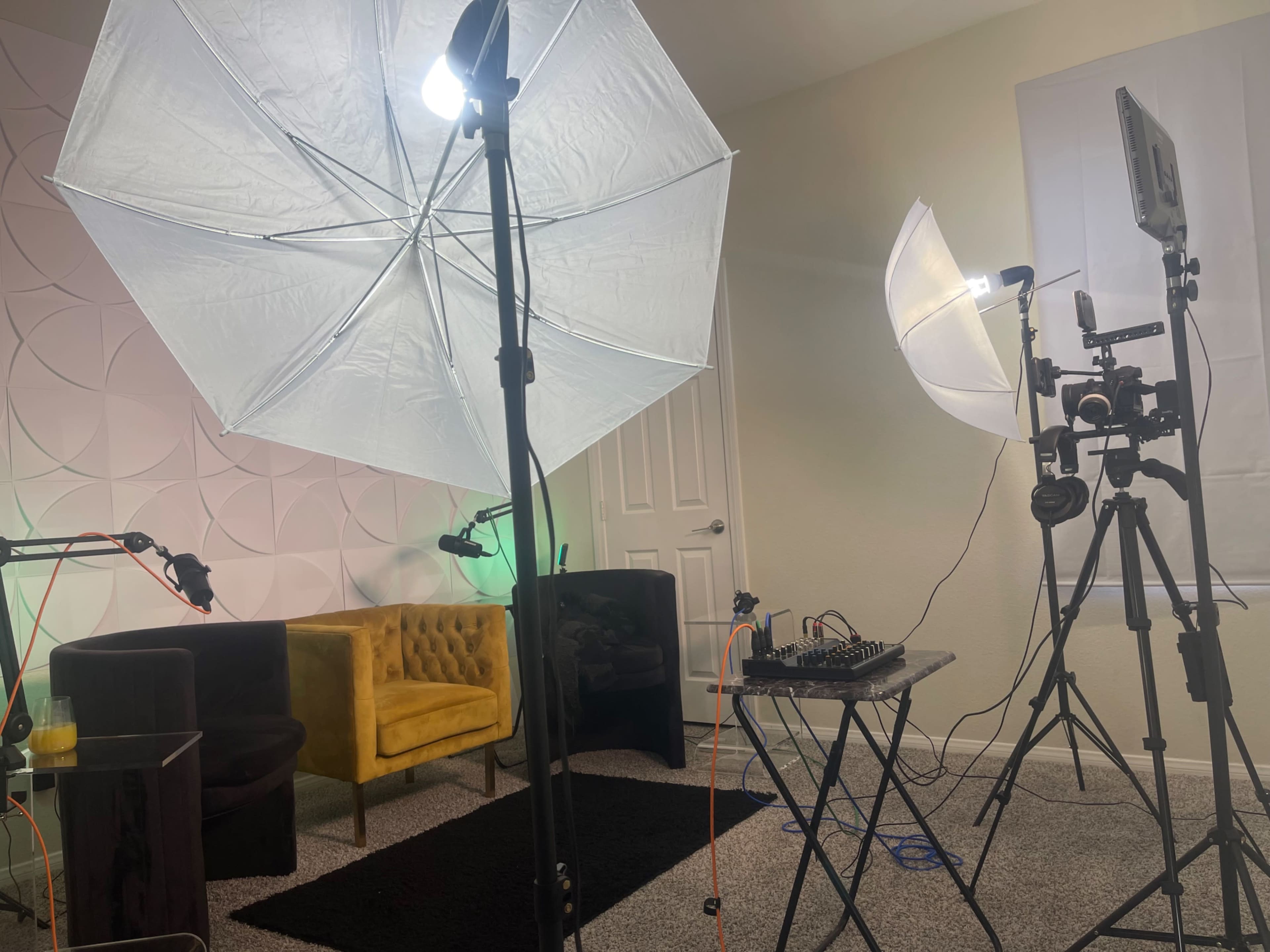 A brightly lit room features two large umbrella lights, a yellow upholstered chair, a black rug, and a table with audio equipment set up for recording.