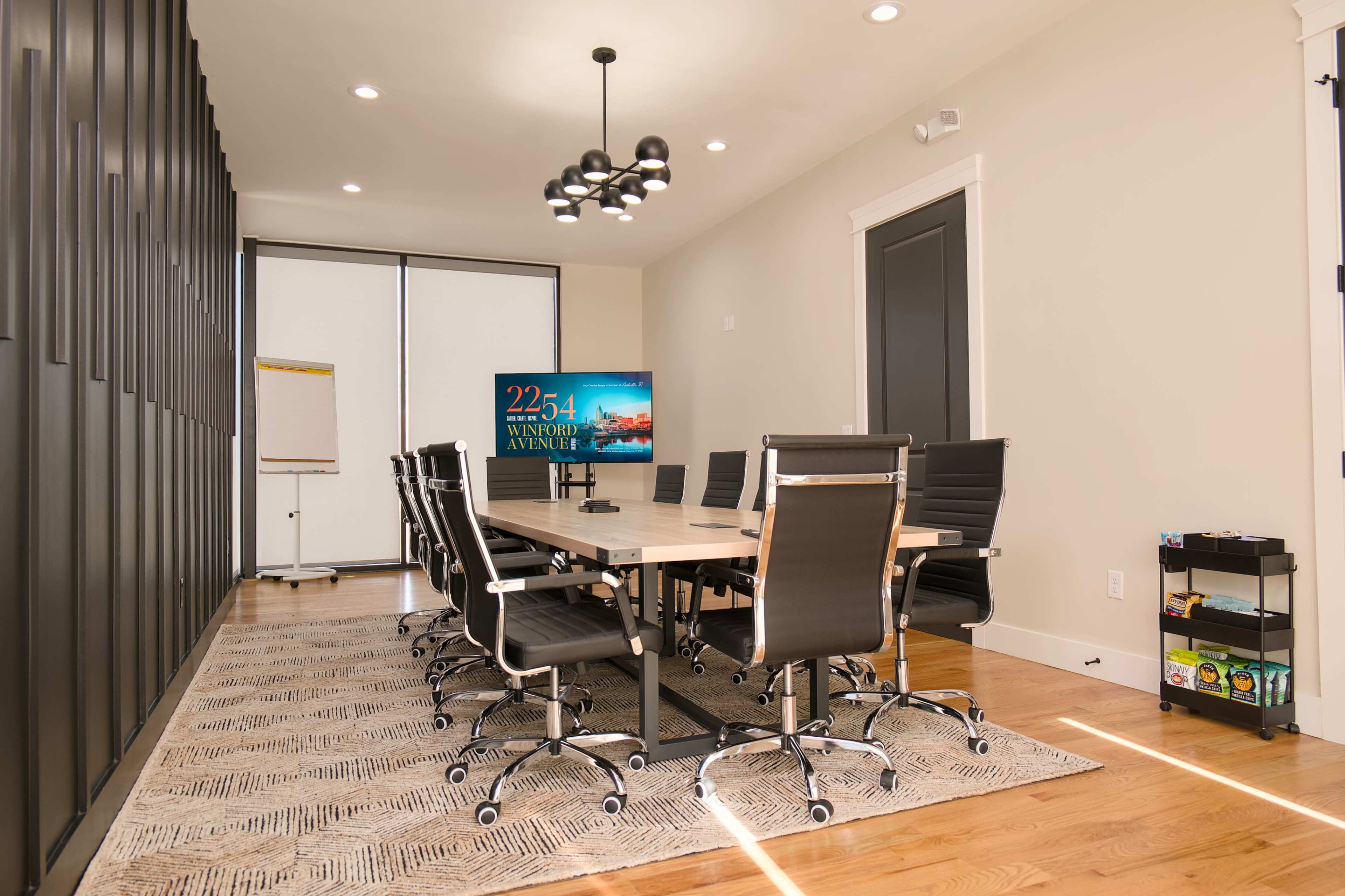 A modern conference room features a large table surrounded by black leather office chairs, a wall-mounted screen displaying information, and large windows with natural light streaming in.