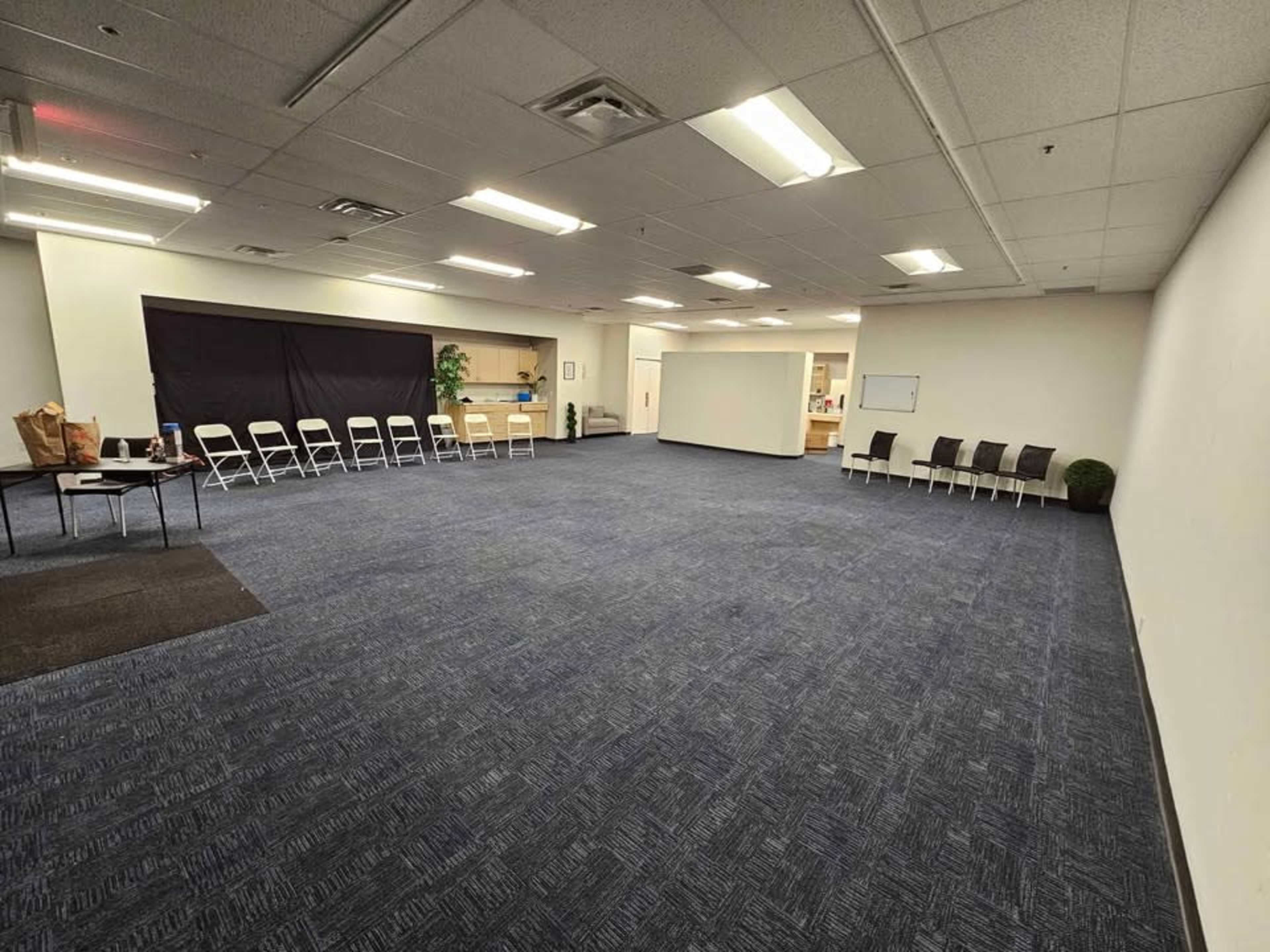 The image shows a large, empty room with blue carpet, a row of white chairs on one side, and a table with bags on it.