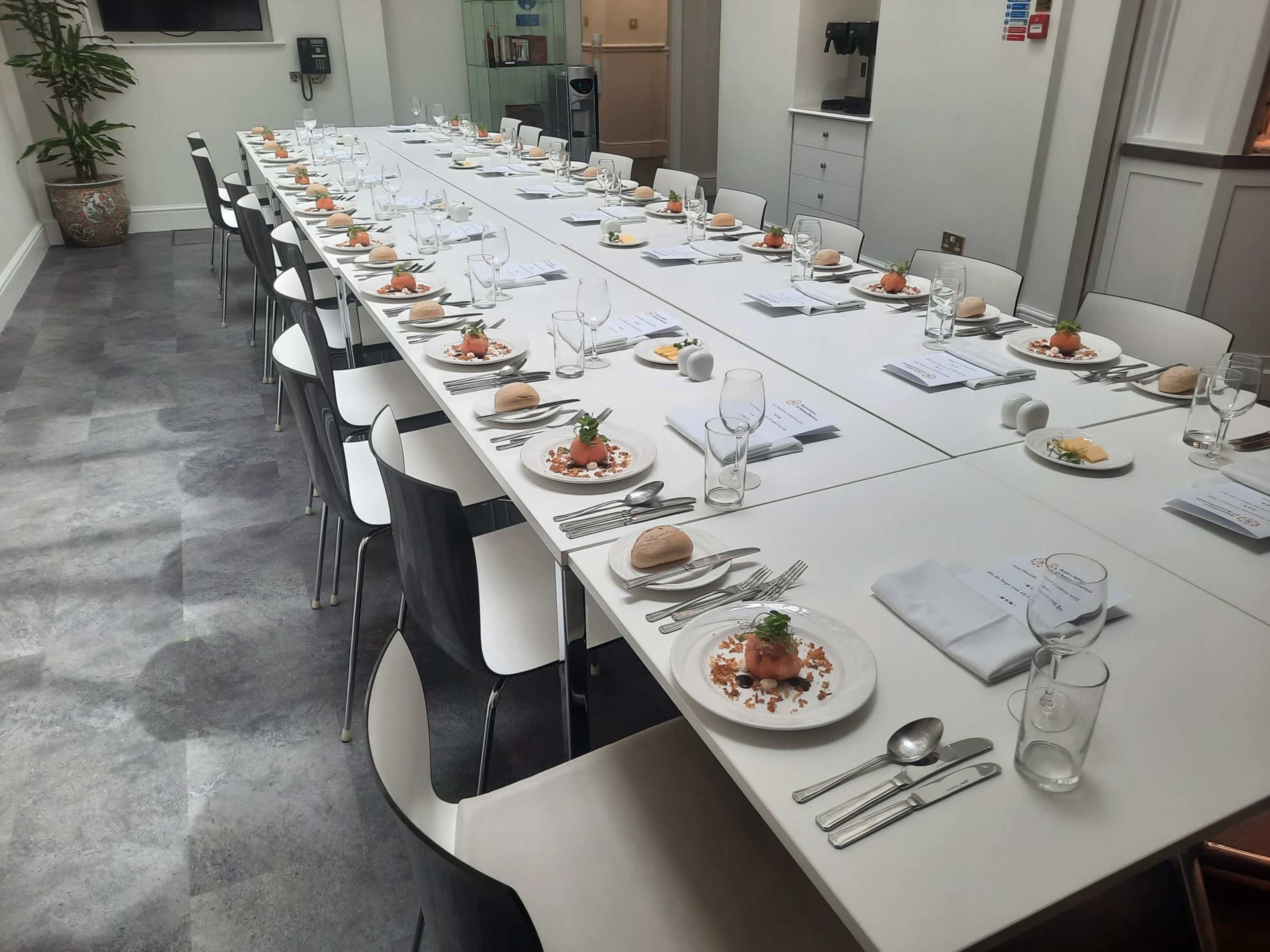 A long dining table is set for a formal meal, featuring neatly arranged plates, cutlery, glasses, and a small bread roll at each place setting.