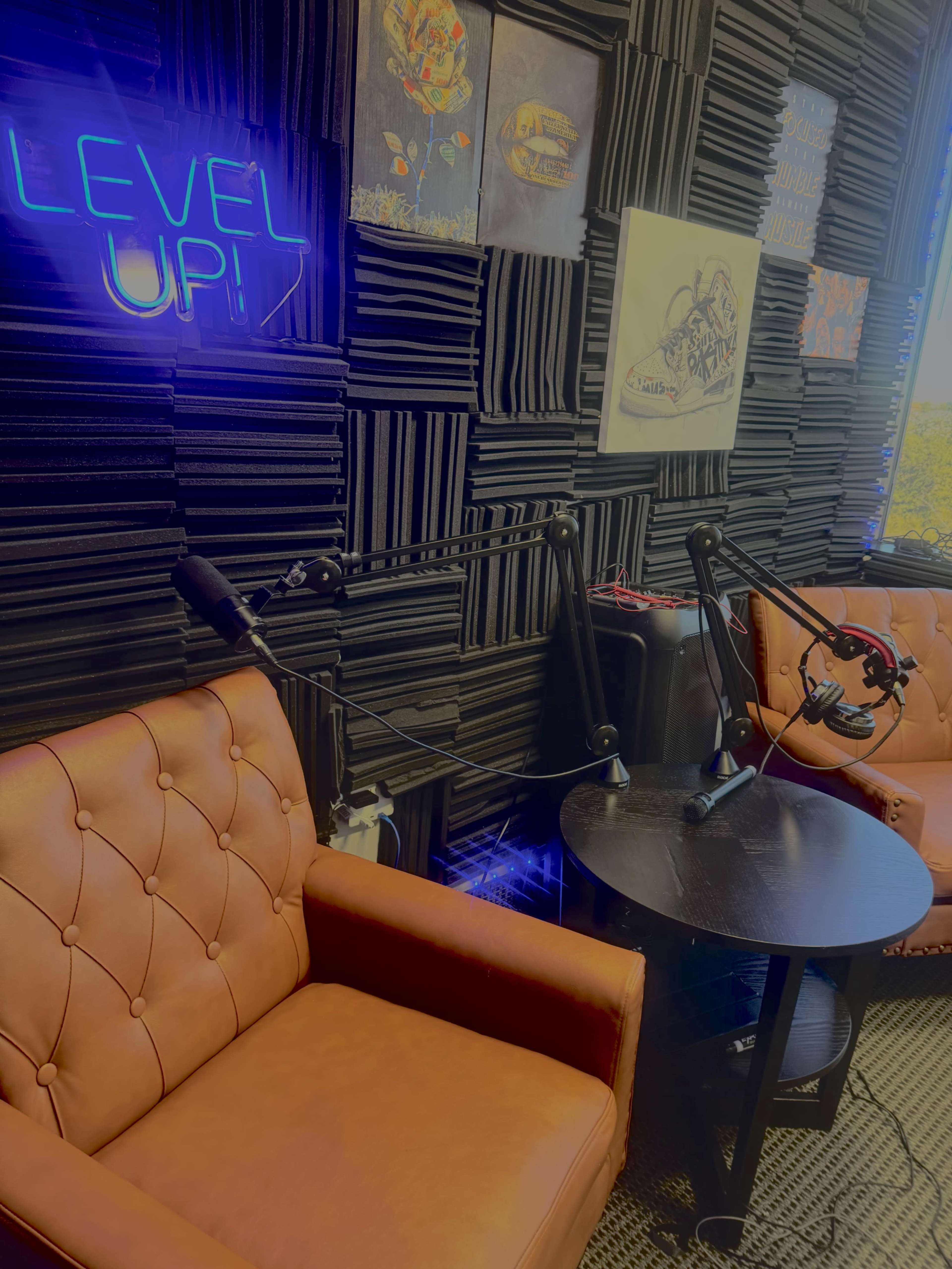 The image shows a cozy recording studio with two brown leather chairs, a small round table, and soundproof wall panels, illuminated by a neon sign reading "LEVEL UP."