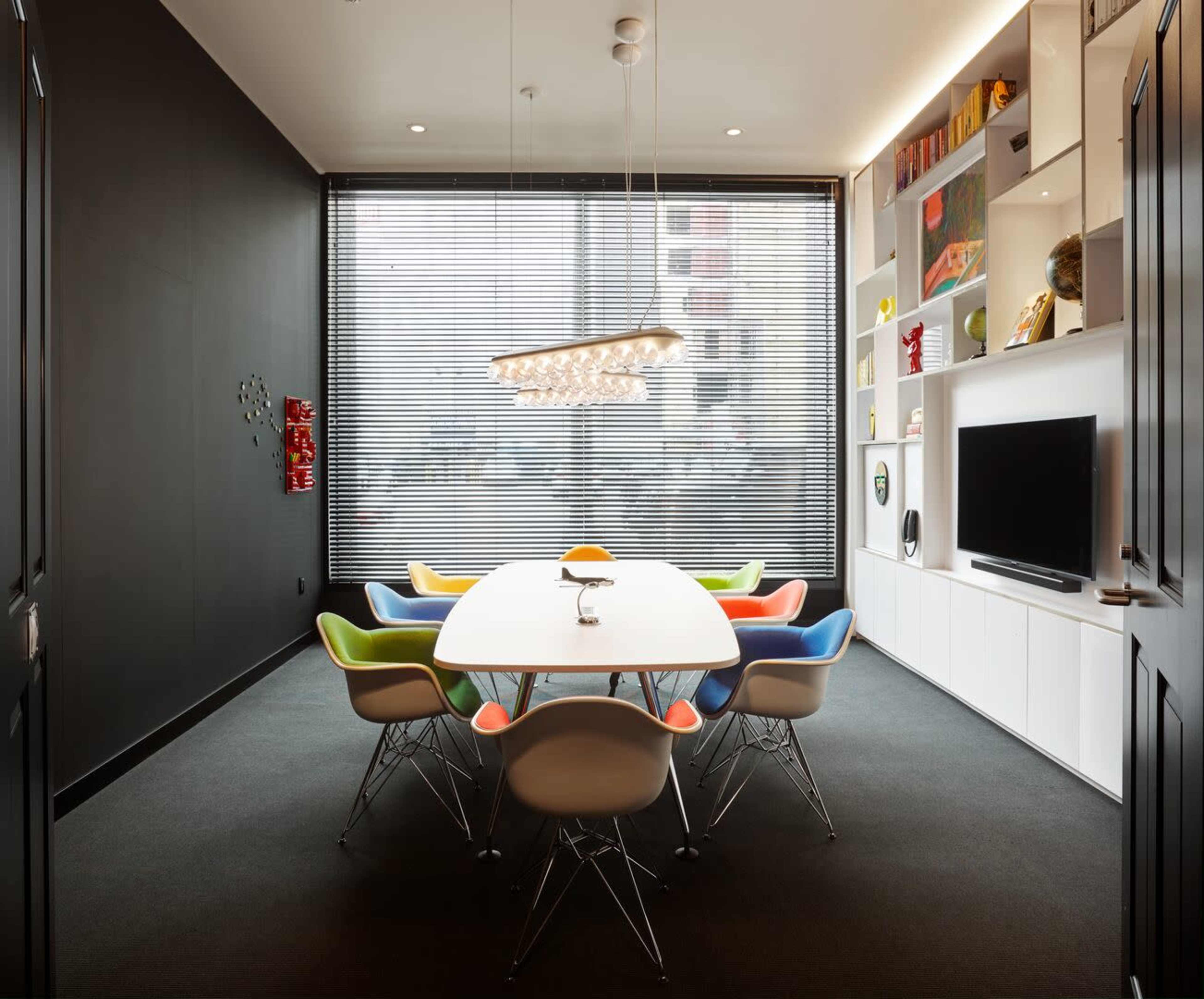 A modern conference room features a long white table surrounded by colorful chairs, with large windows that have slatted blinds.