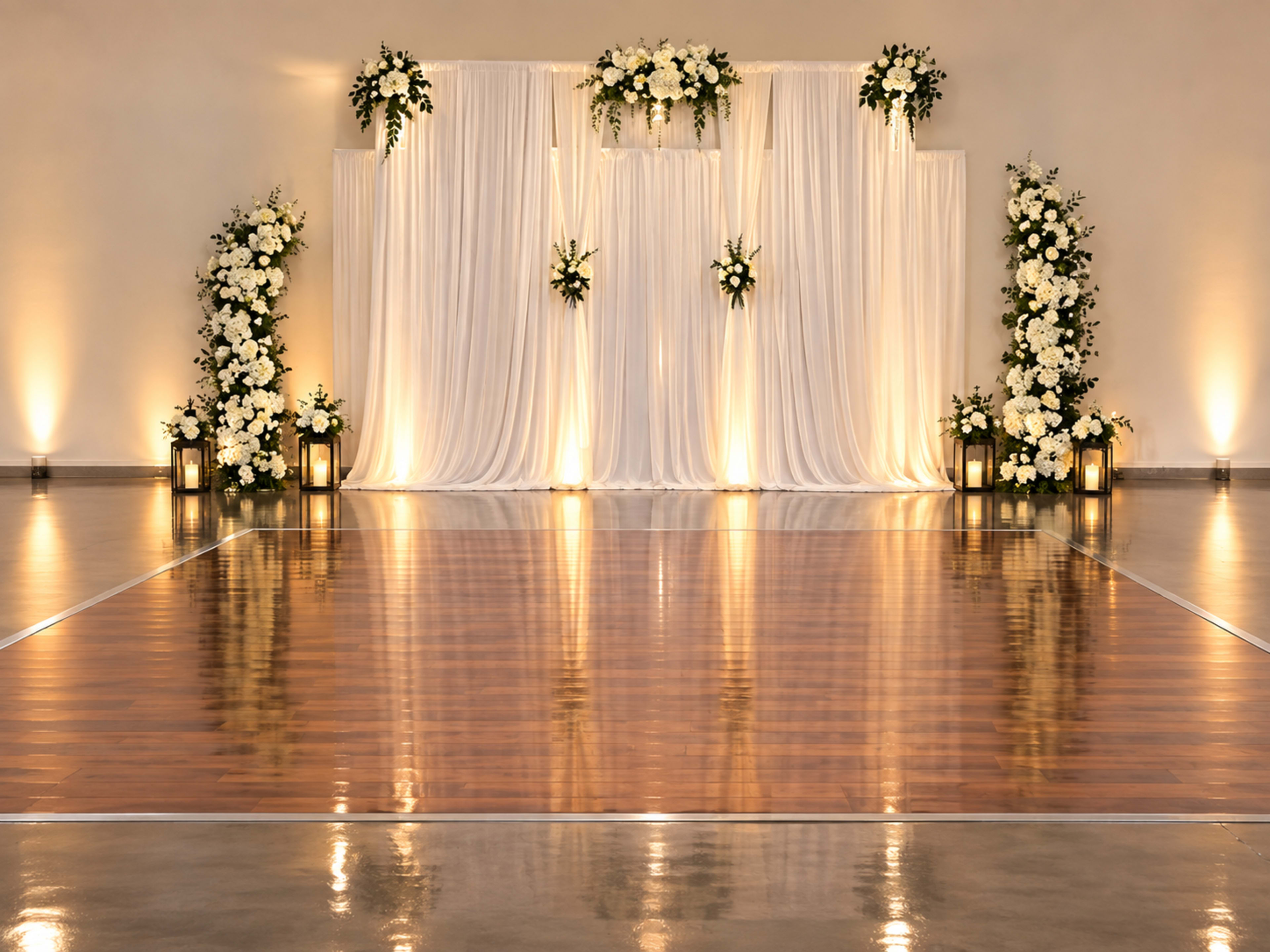 The image shows a decorated stage with white drapes and floral arrangements, illuminated by soft lighting, set against a polished wooden dance floor.