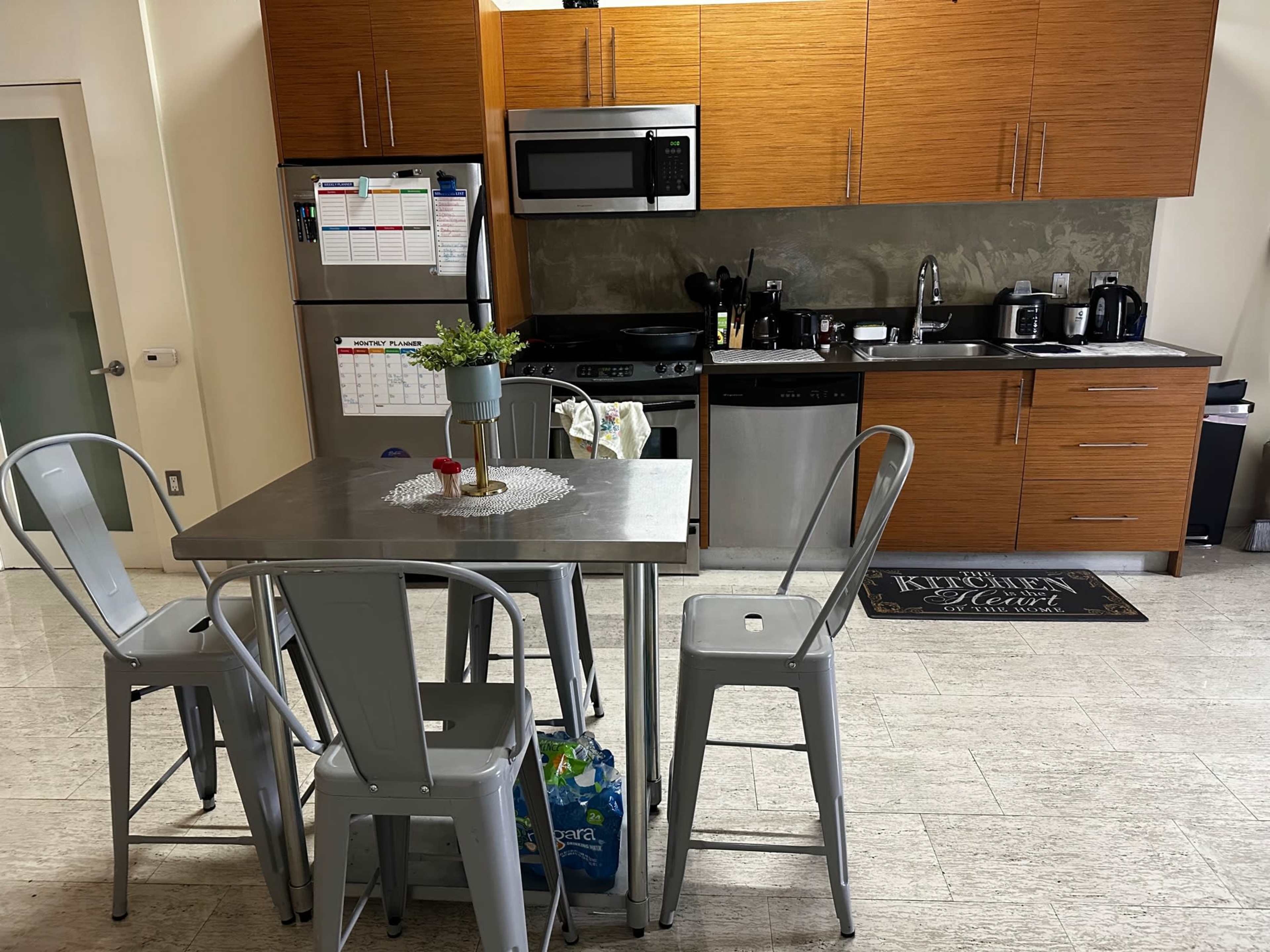 A modern kitchen with stainless steel appliances, wooden cabinets, a table surrounded by metal chairs, and a small plant centerpiece.