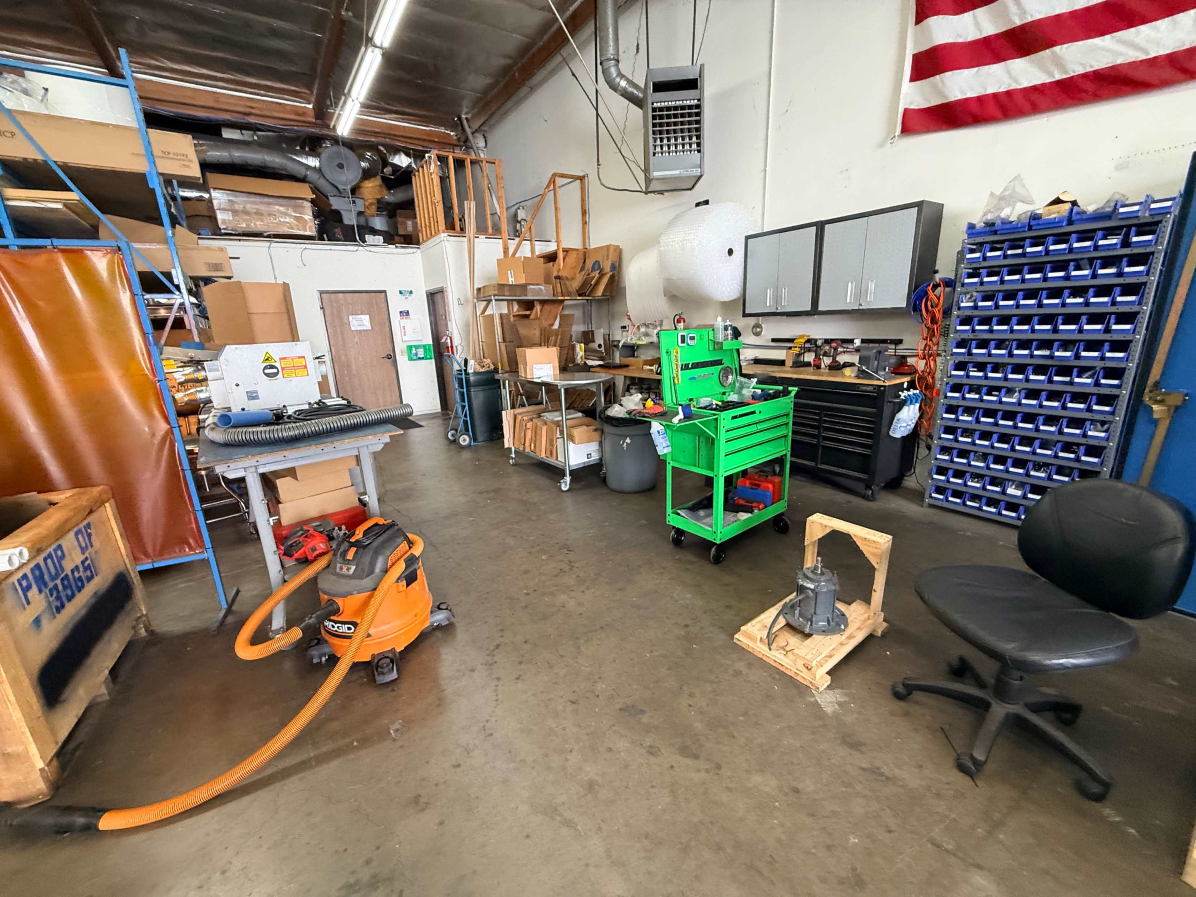 The image shows a workshop with various tools, equipment, and storage areas, including a workbench, a vacuum, and a chair.