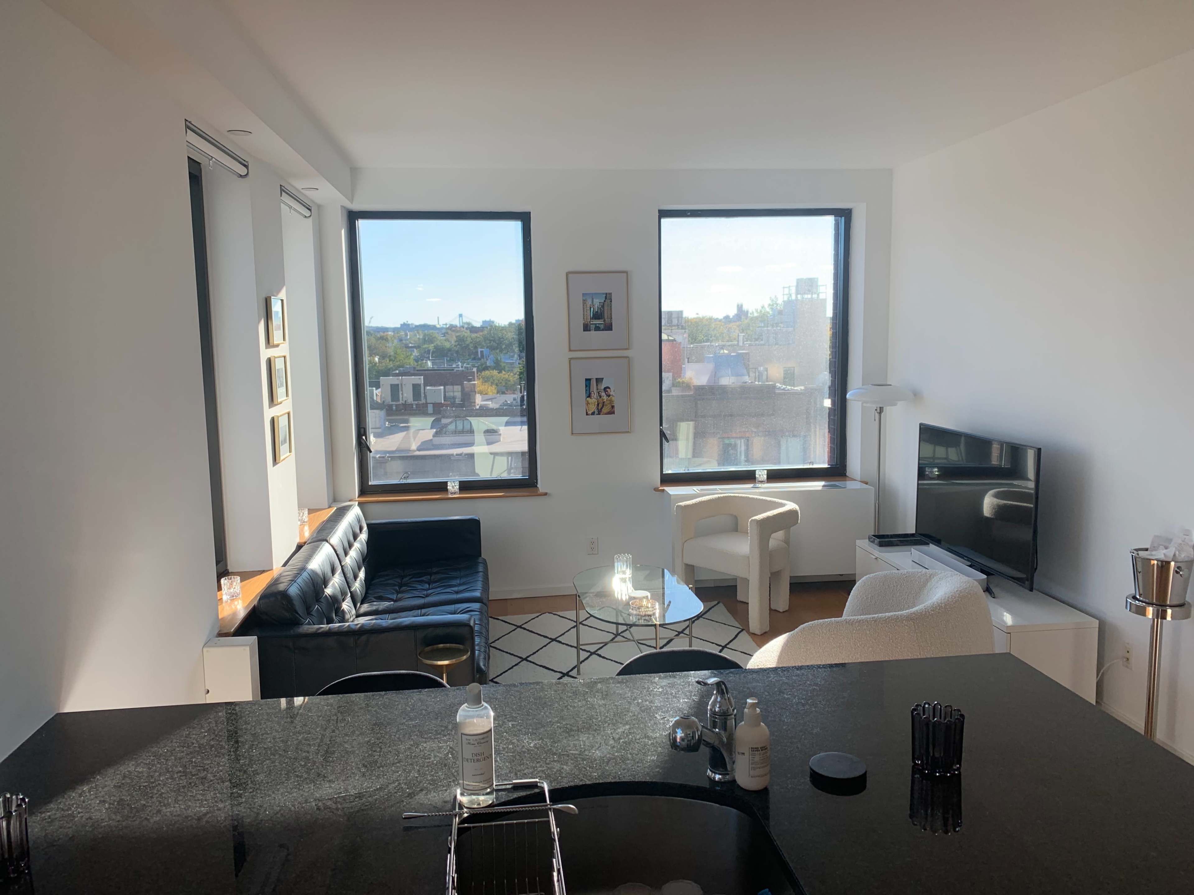 A modern living room features a black leather sofa, a white armchair, a glass coffee table, and a television, with large windows displaying a cityscape.