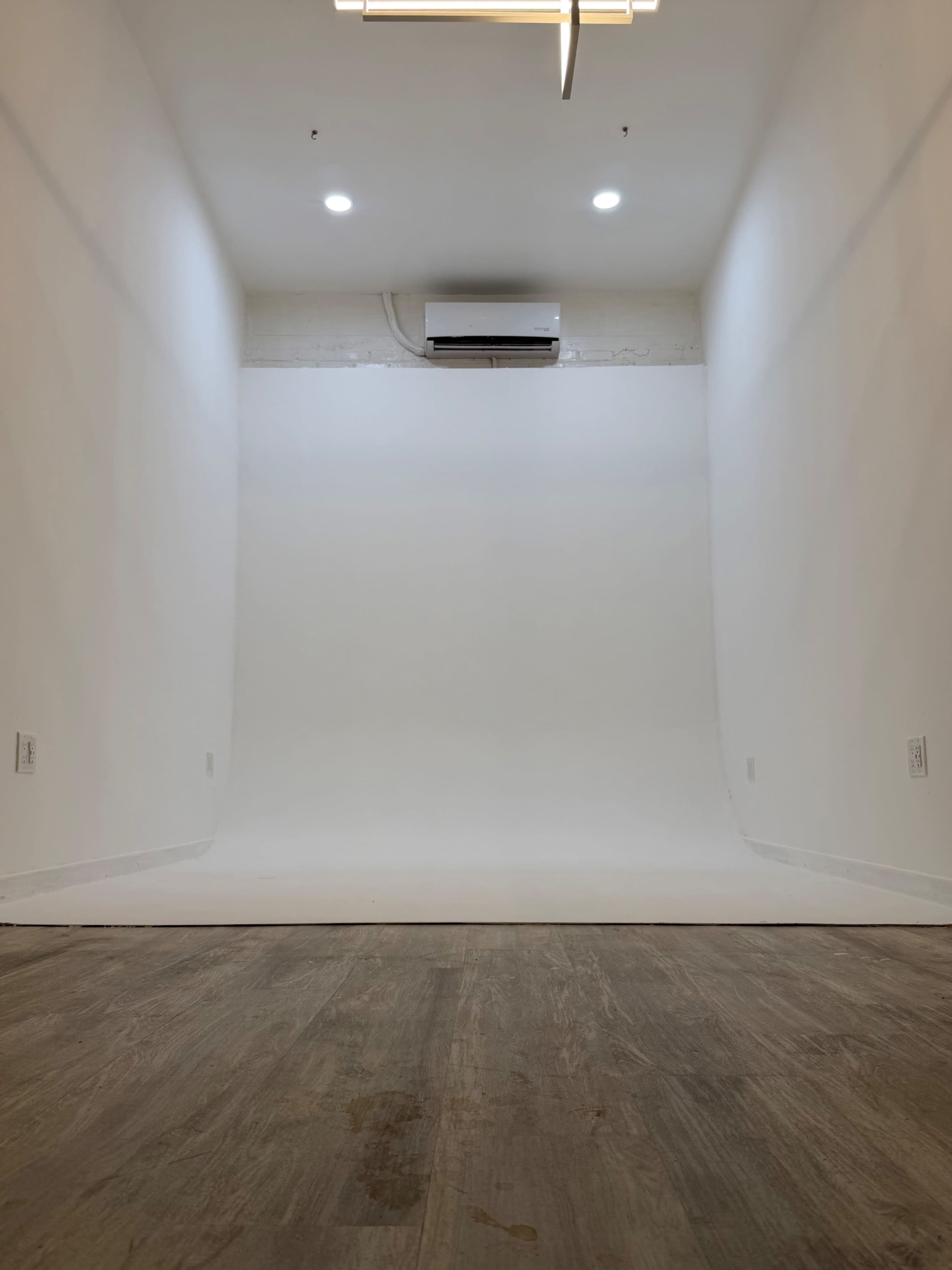 The image shows a minimalist, empty room with a seamless white backdrop and wooden flooring.