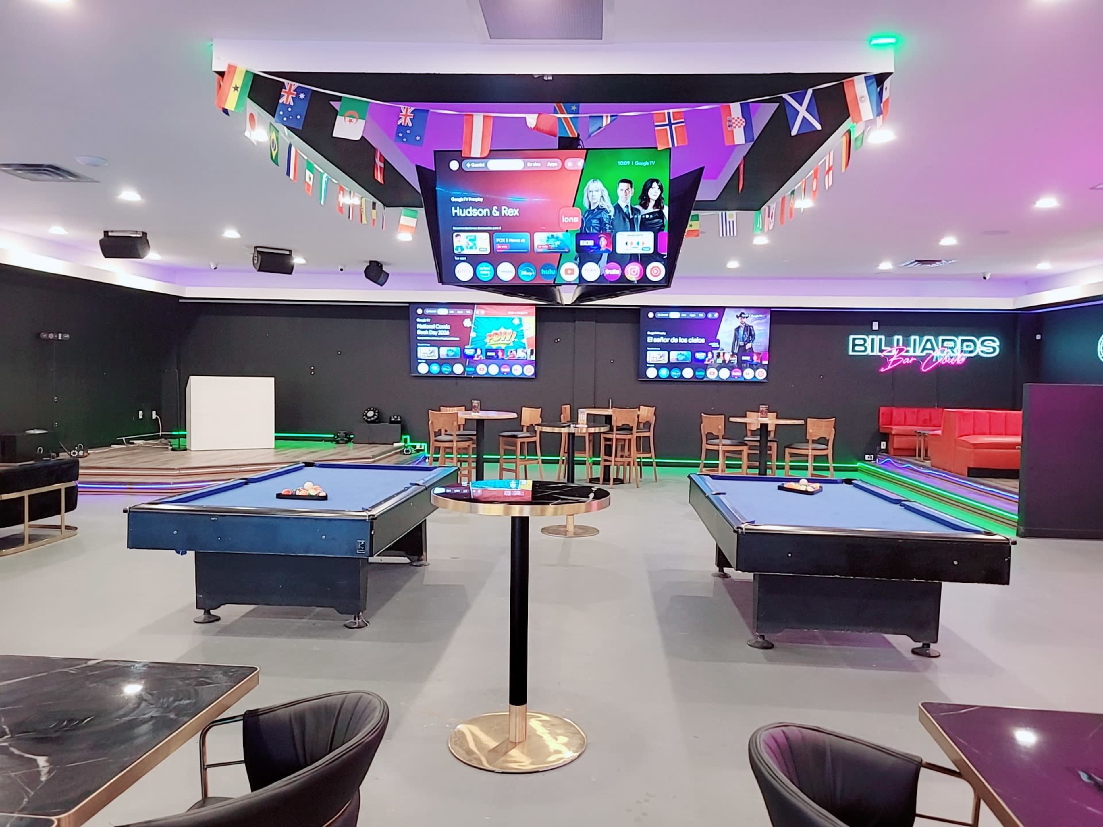 The image shows a modern billiards lounge with two pool tables, seating areas, and multiple screens displaying game statistics, surrounded by flags from various countries.