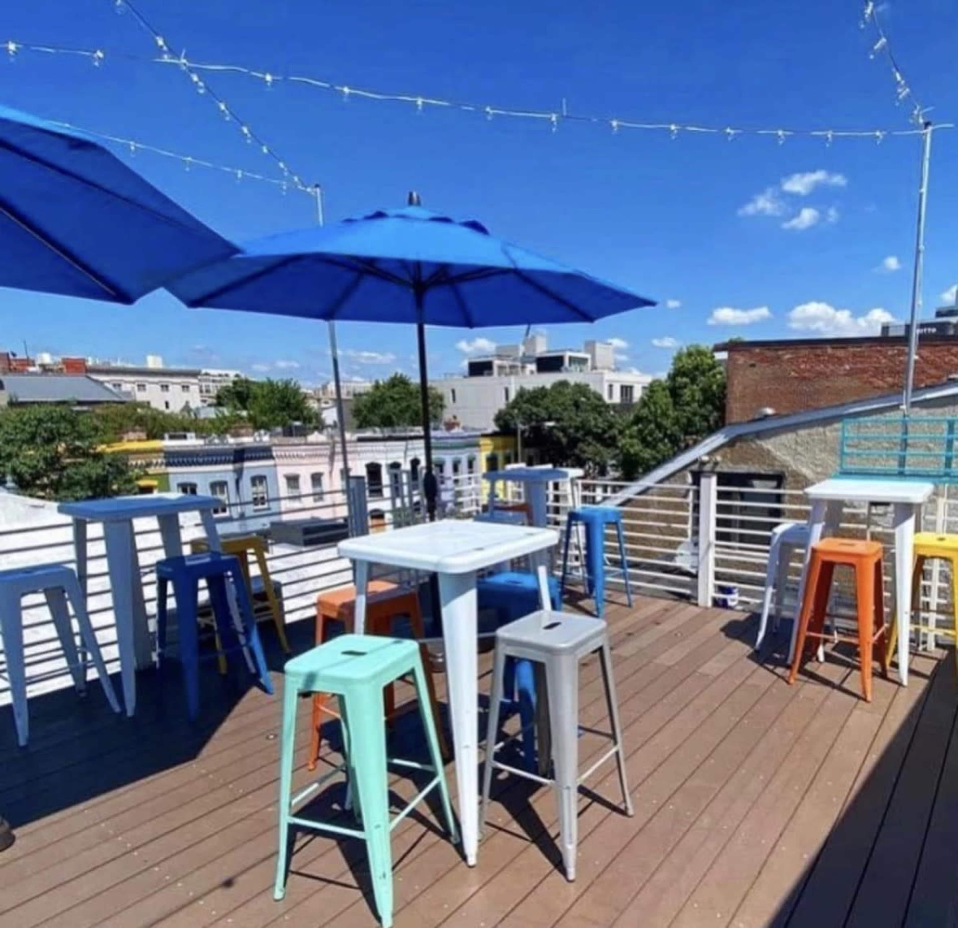 The image shows a rooftop terrace with several brightly colored stools and tables under blue umbrellas, surrounded by buildings and a clear sky.