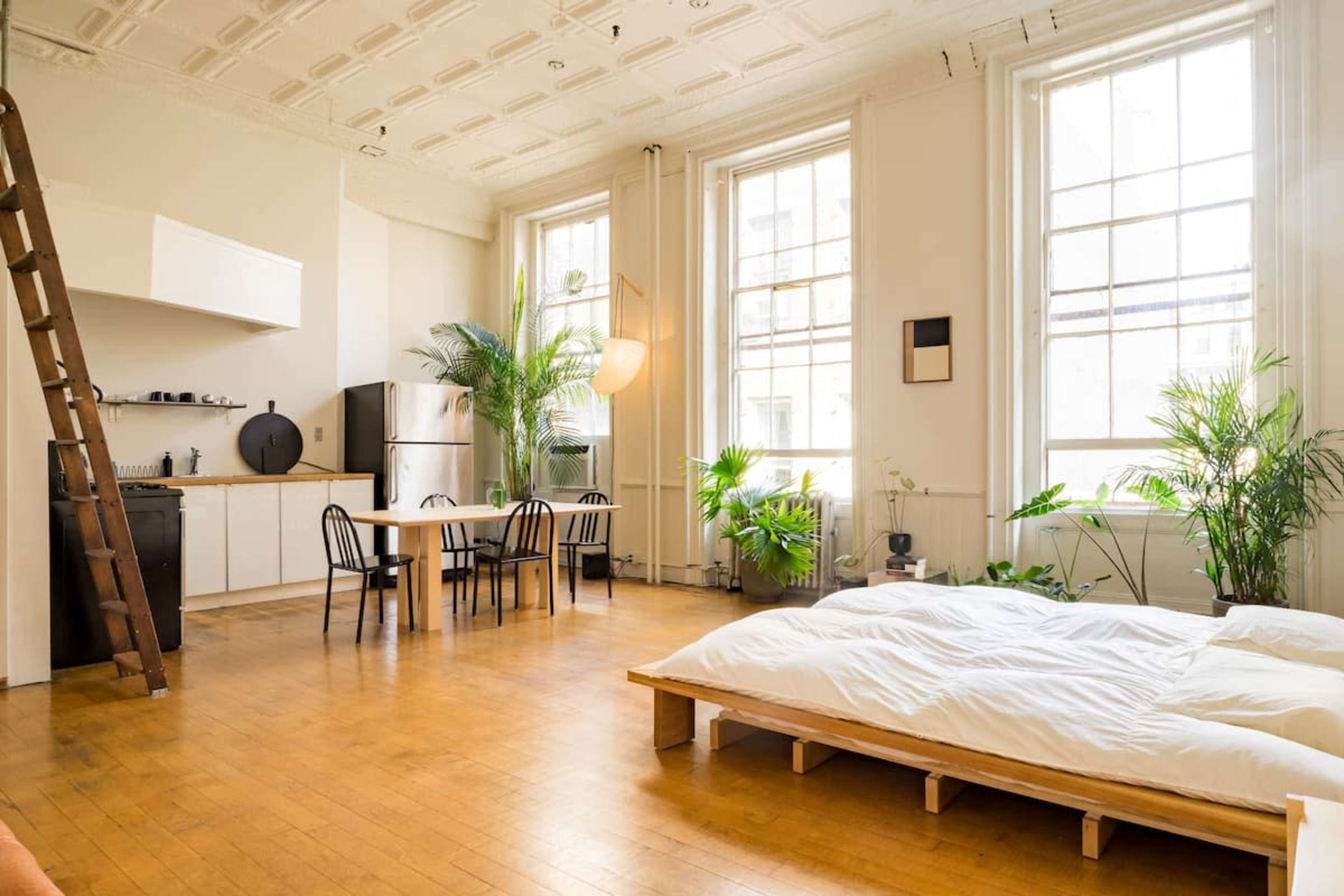 A bright, minimalist studio apartment with a kitchen area, dining table, and a bed on a wooden platform surrounded by plants.