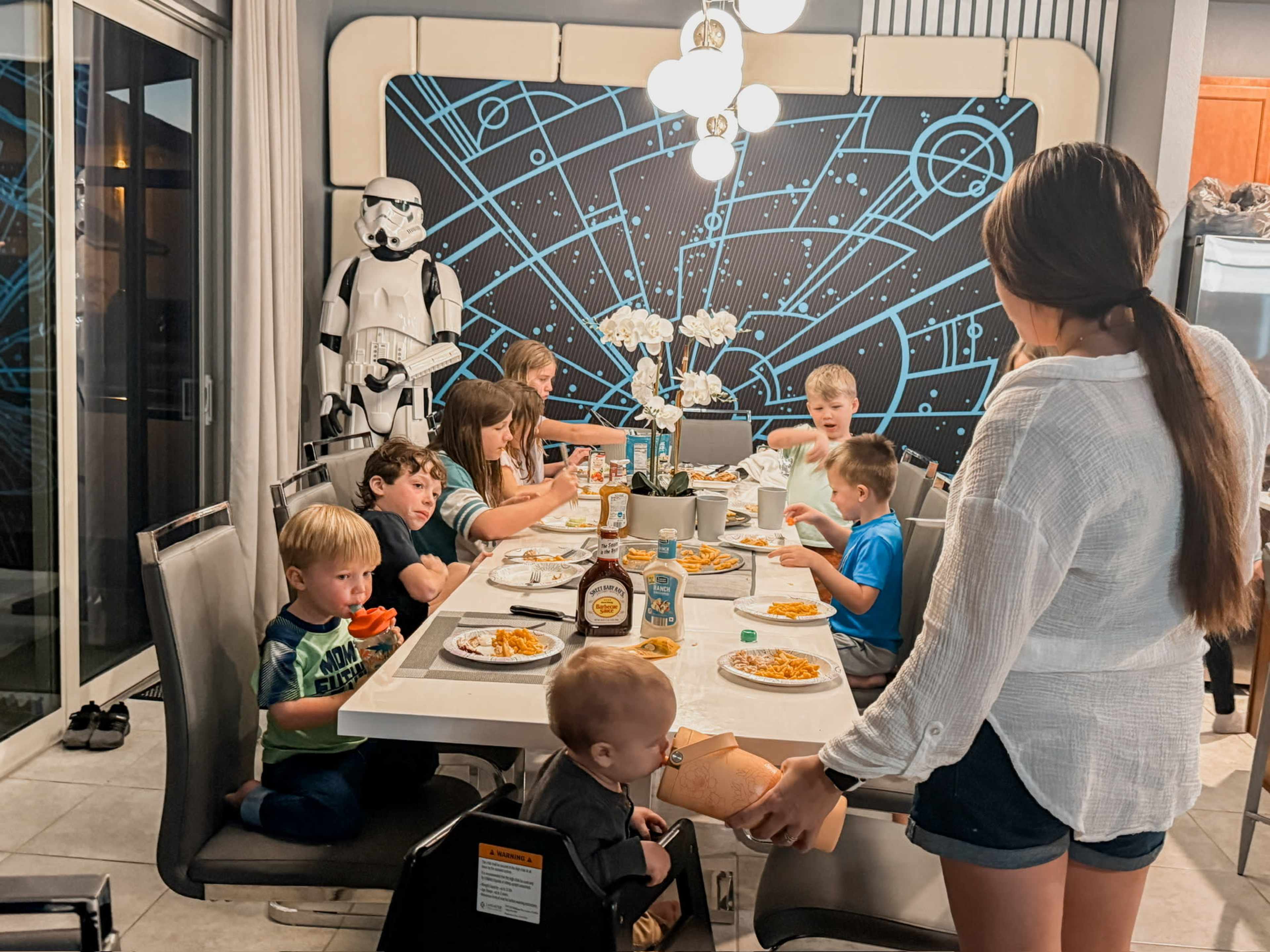 A group of children and an adult sit around a dining table enjoying dinner, with a Star Wars-themed wall design and a stormtrooper figure in the background.