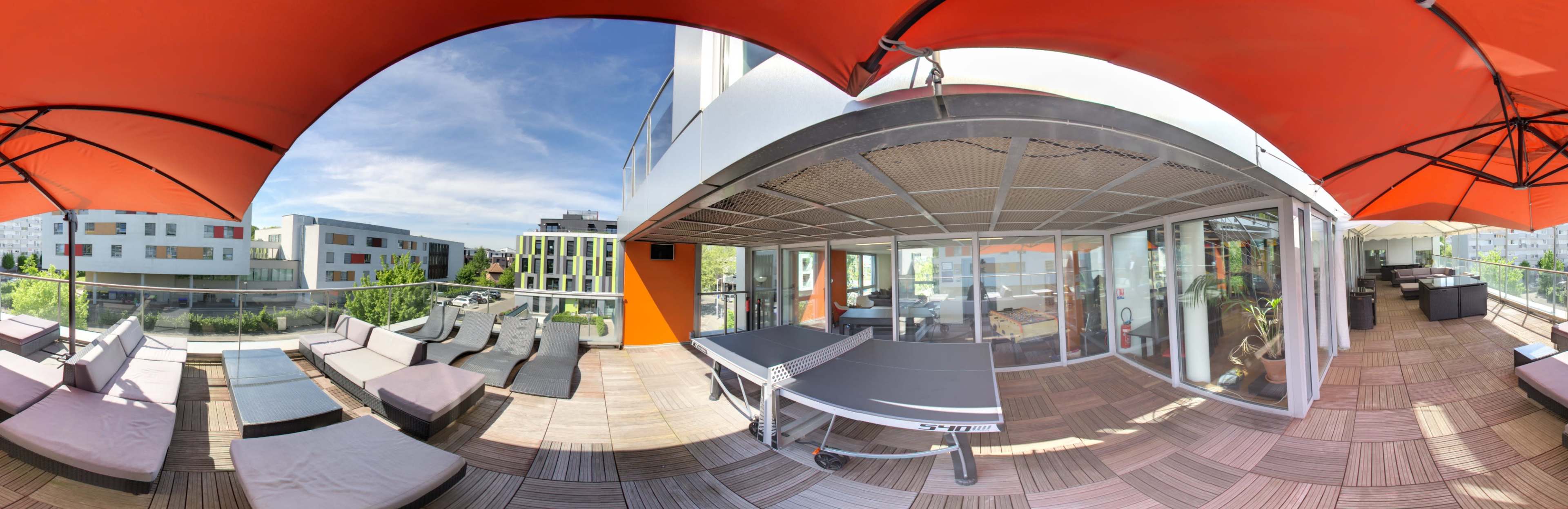 A panoramic view of a rooftop terrace featuring lounge chairs, a ping pong table, and a backdrop of modern buildings under a clear sky.