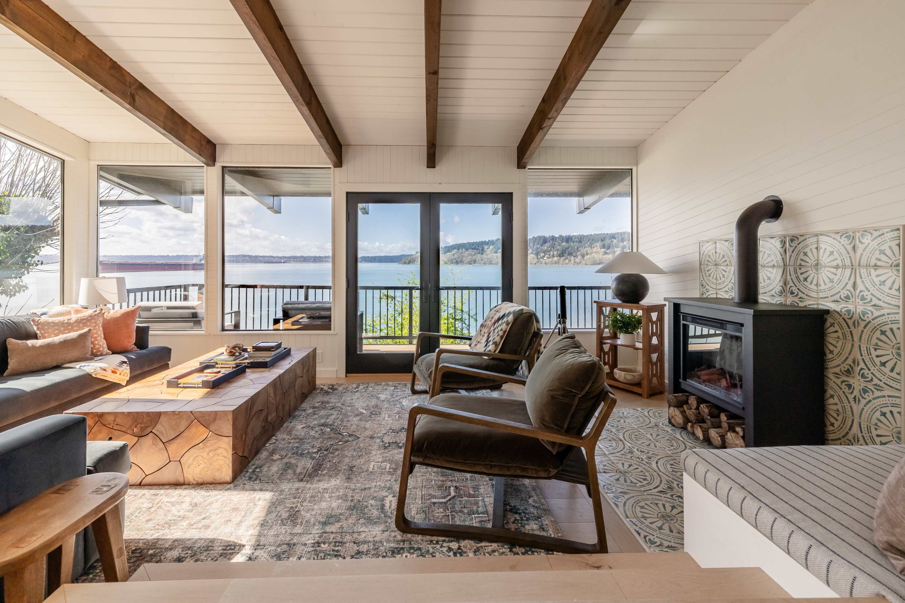 A spacious living area features large windows overlooking a body of water, with comfortable seating, a wooden coffee table, and a fireplace.