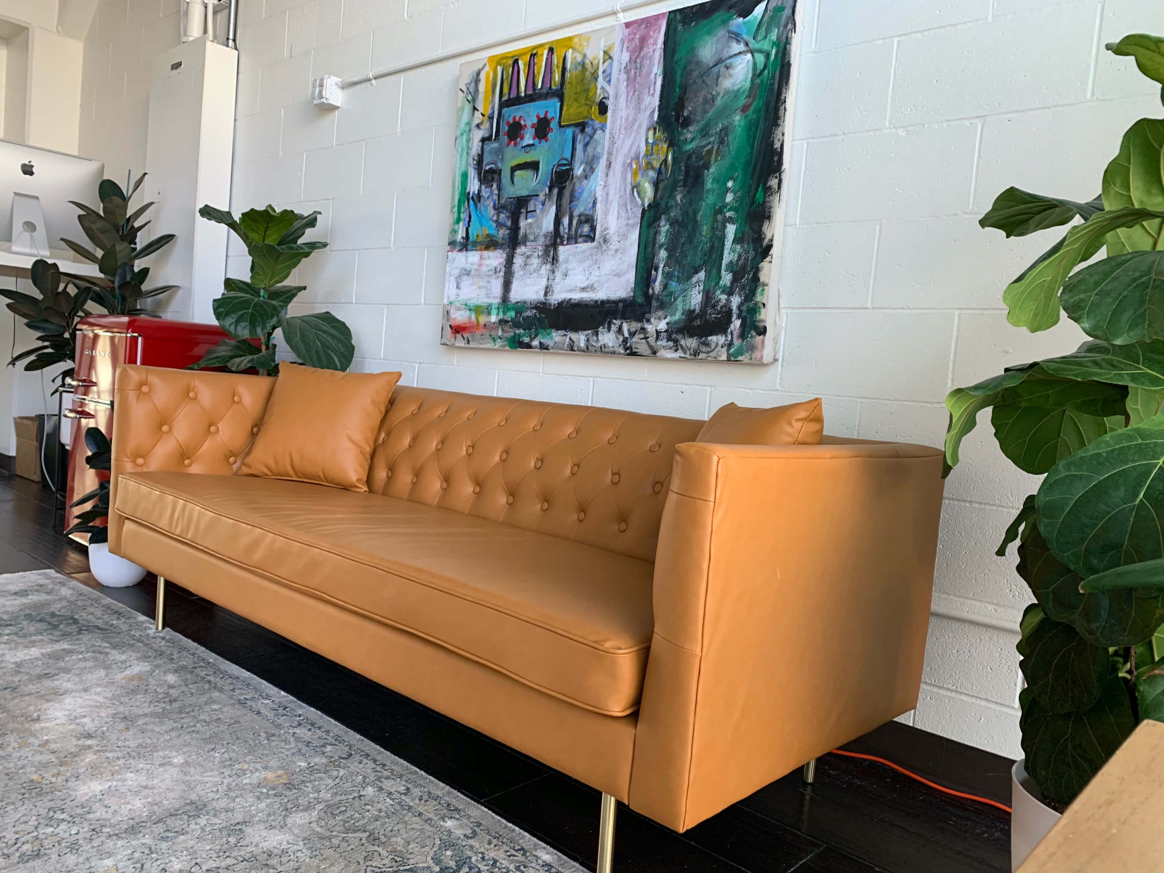 A tan tufted leather sofa is positioned against a white wall, complemented by a colorful abstract painting and large green plants nearby.