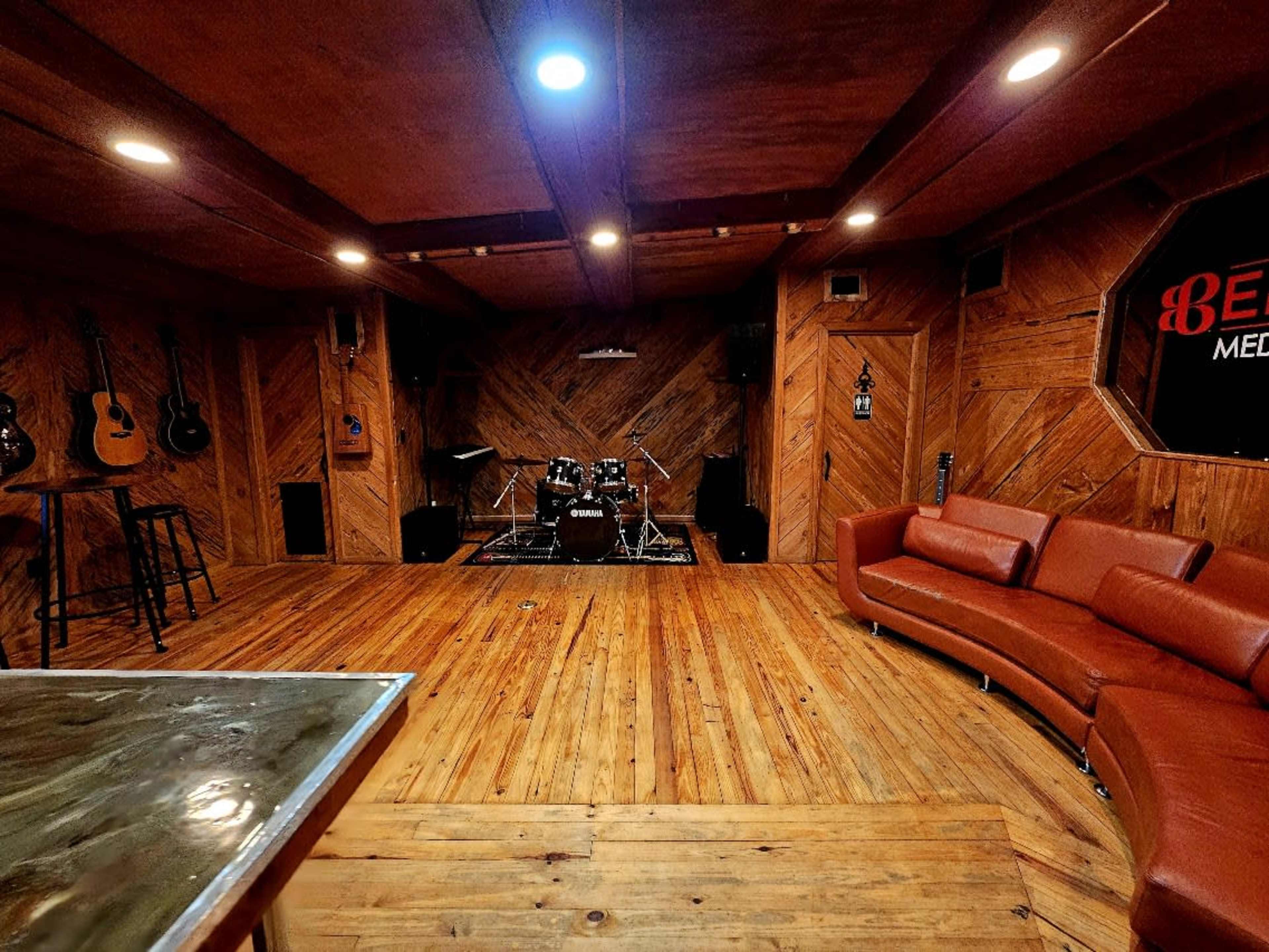 The image shows a music rehearsal space featuring a wooden floor, walls lined with guitars, a drum set, and a brown leather couch in the corner.