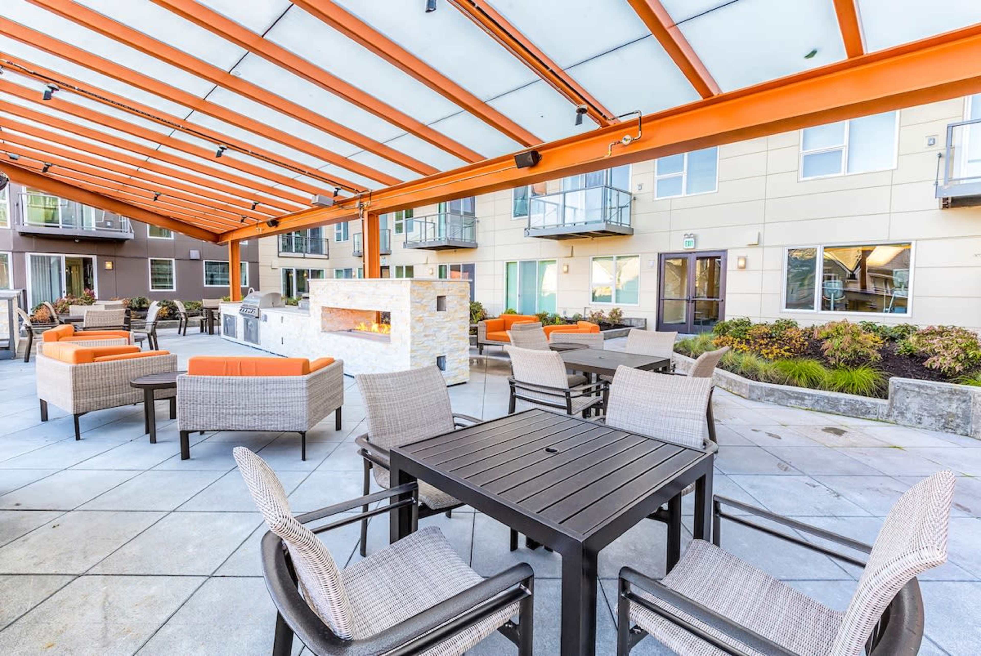 The image shows a spacious outdoor patio area furnished with tables, chairs, and couches, all sheltered under a metal frame with an overhead canopy.