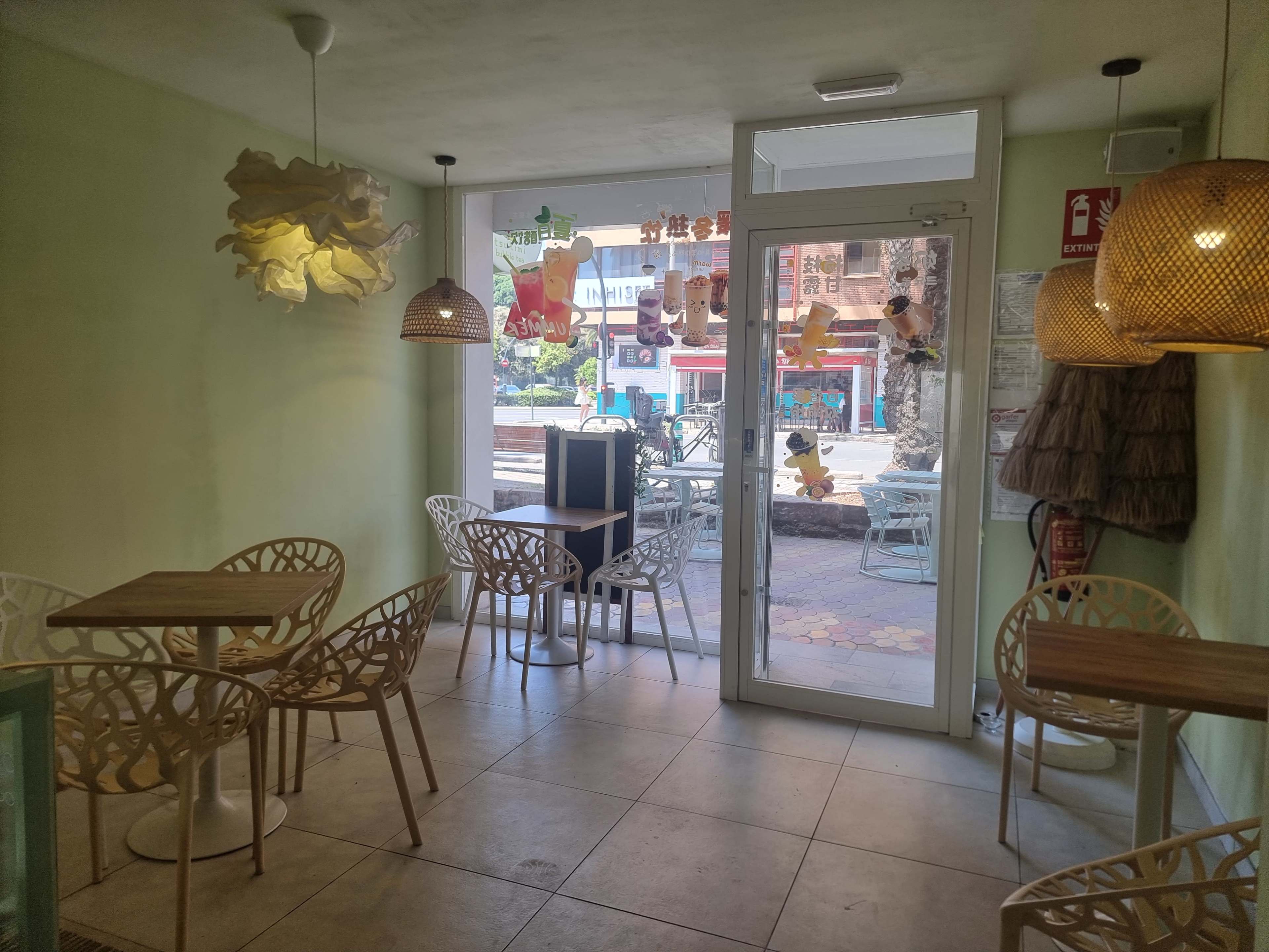 A small café features light-colored walls, several wooden tables, and decorative hanging lights, with a view of an outdoor seating area through a glass door.