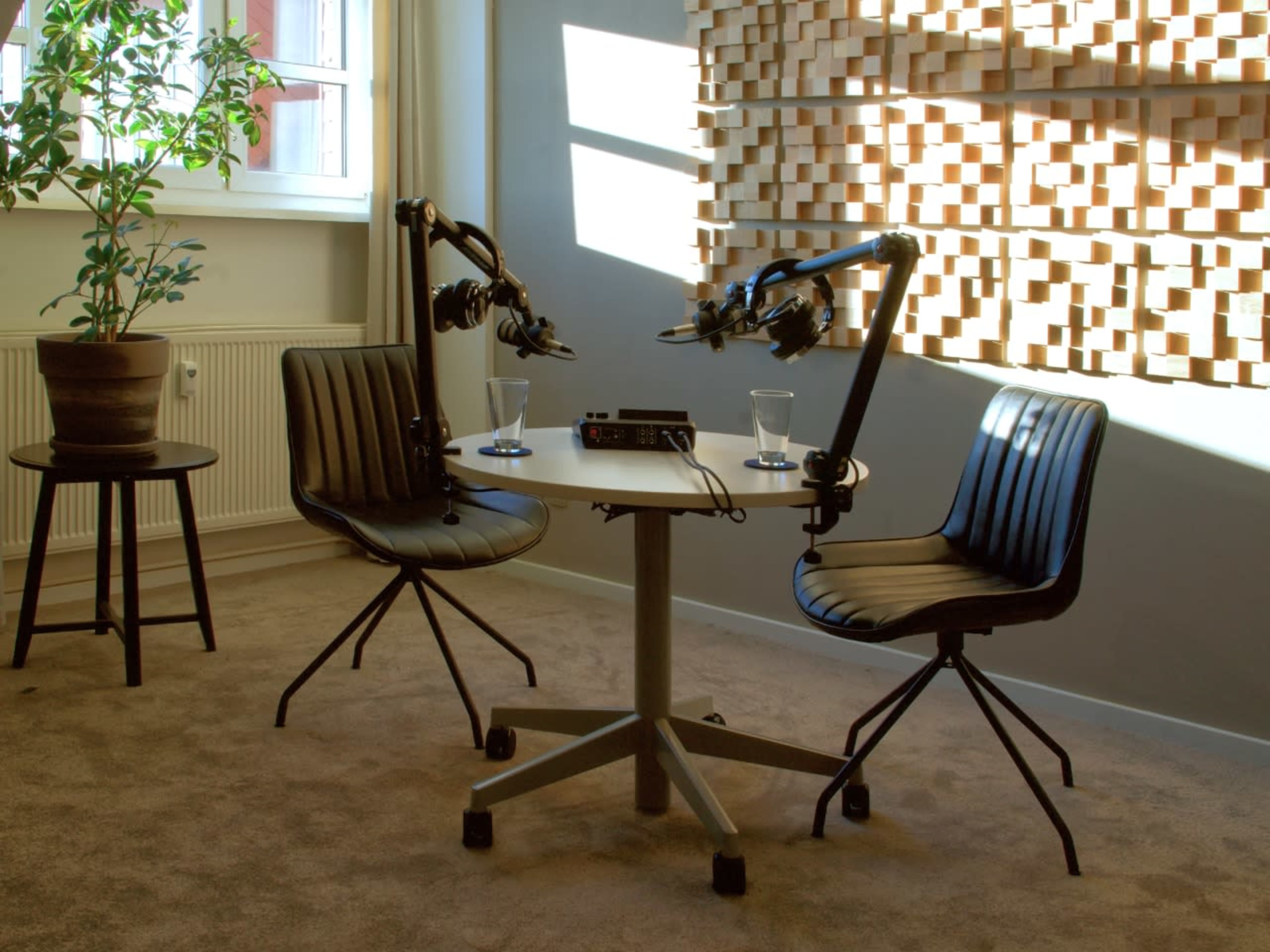 The image shows a podcast studio setup with two chairs, a small table, and microphone arms, illuminated by natural light from a window.