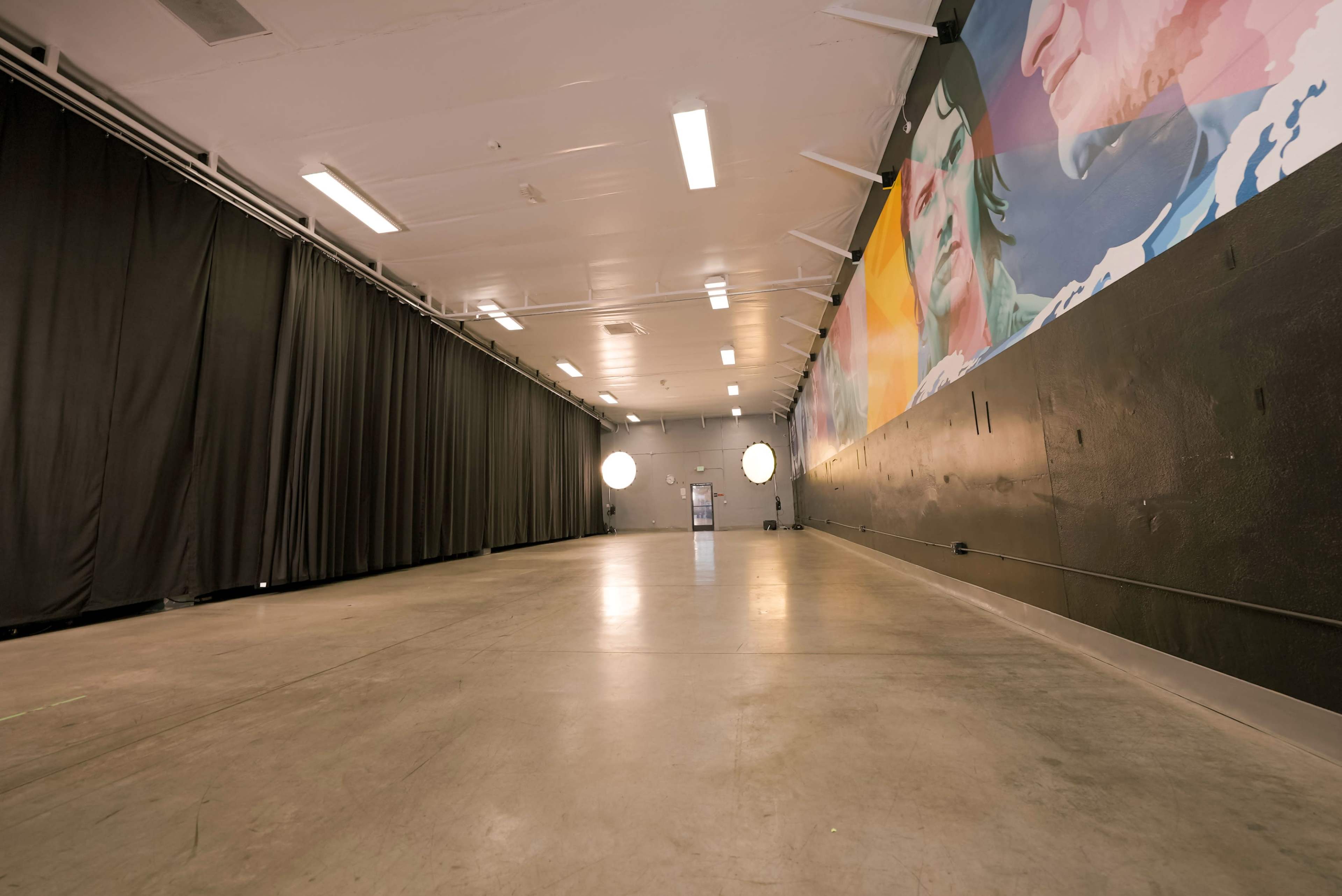 A long, empty interior space with a mural on one wall and black curtains lining the opposite side.