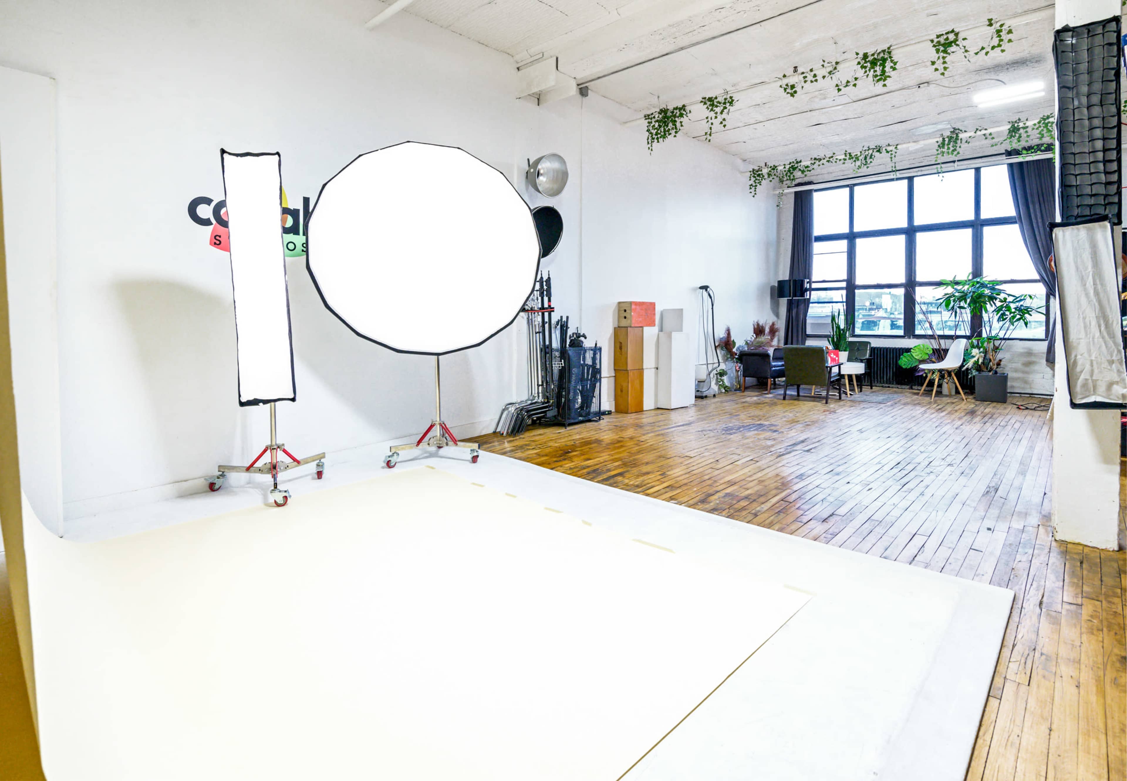 The image shows a spacious photography studio with wooden flooring, large windows, and various lighting equipment setup.